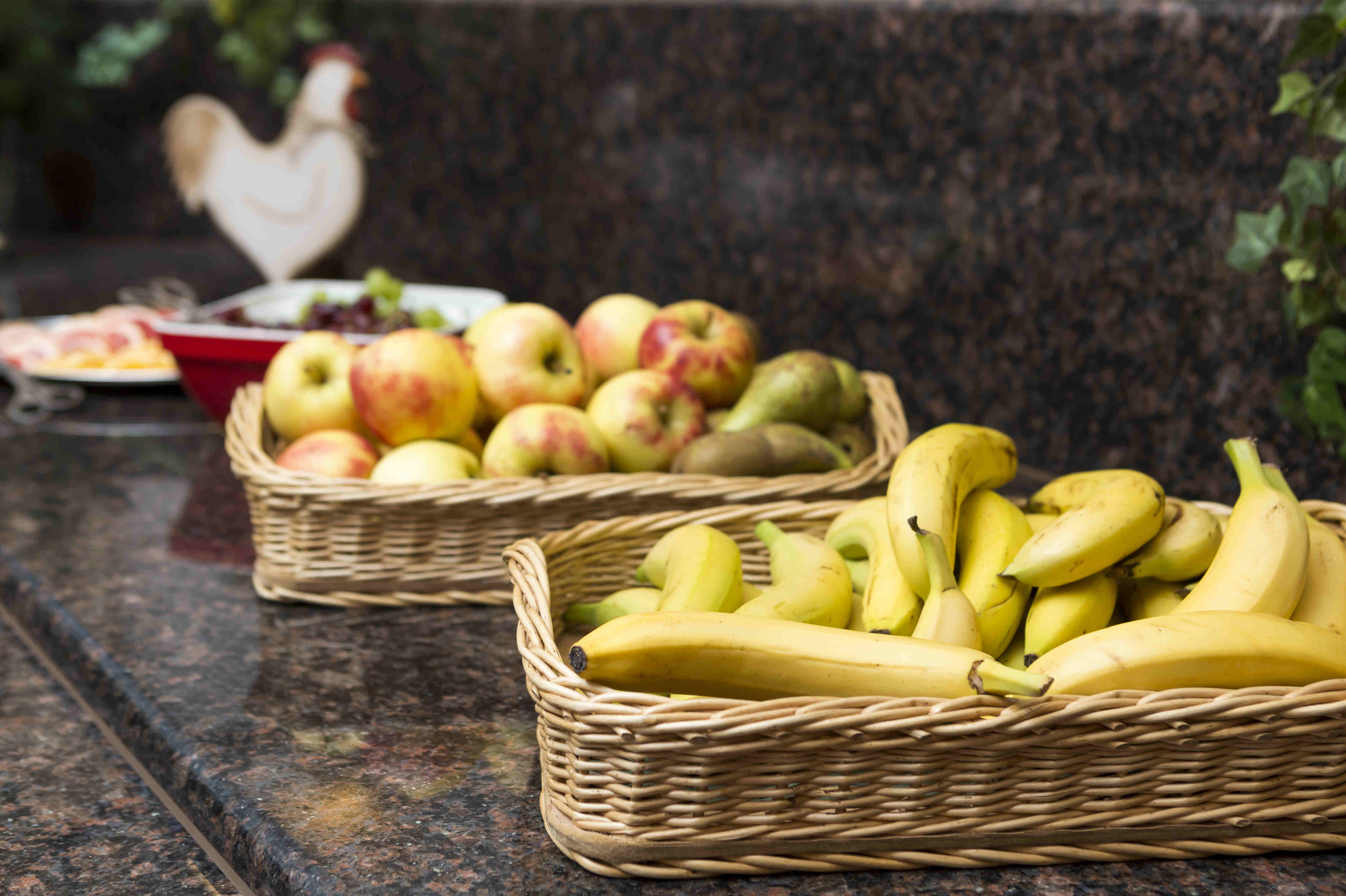 Sportpark Rabenberg: Obst beim Frühstücksbuffet Auf einer glänzenden, dunklen Granit-Arbeitsplatte stehen zwei geflochtene Obstkörbe aus hellem Naturmaterial. Der vordere Korb ist prall gefüllt mit gelben Bananen. Direkt dahinter steht ein zweiter Korb, in dem eine bunte Mischung aus Äpfeln, Birnen und möglicherweise Kiwis liegt. Die Früchte sind sorgfältig arrangiert und wirken frisch und einladend.