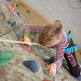 Sportpark Rabenberg: Kletterwand Das Bild zeigt ein Kind beim Klettern an einer Kletterwand. Das Kind ist von oben fotografiert, so dass man es von Kopf bis Fuß sehen kann, während es die Wand erklimmt. Es ist ein junges Mädchen, dessen blondes Haar zu einem kleinen Pferdeschwanz zusammengebunden ist. Sie trägt ein langärmliges rot-weiß gestreiftes Oberteil, schwarze Leggings und einen blauen Klettergurt. Ein Sicherungsseil ist an dem Klettergurt des Kindes befestigt und verläuft nach oben und aus dem Bild heraus.