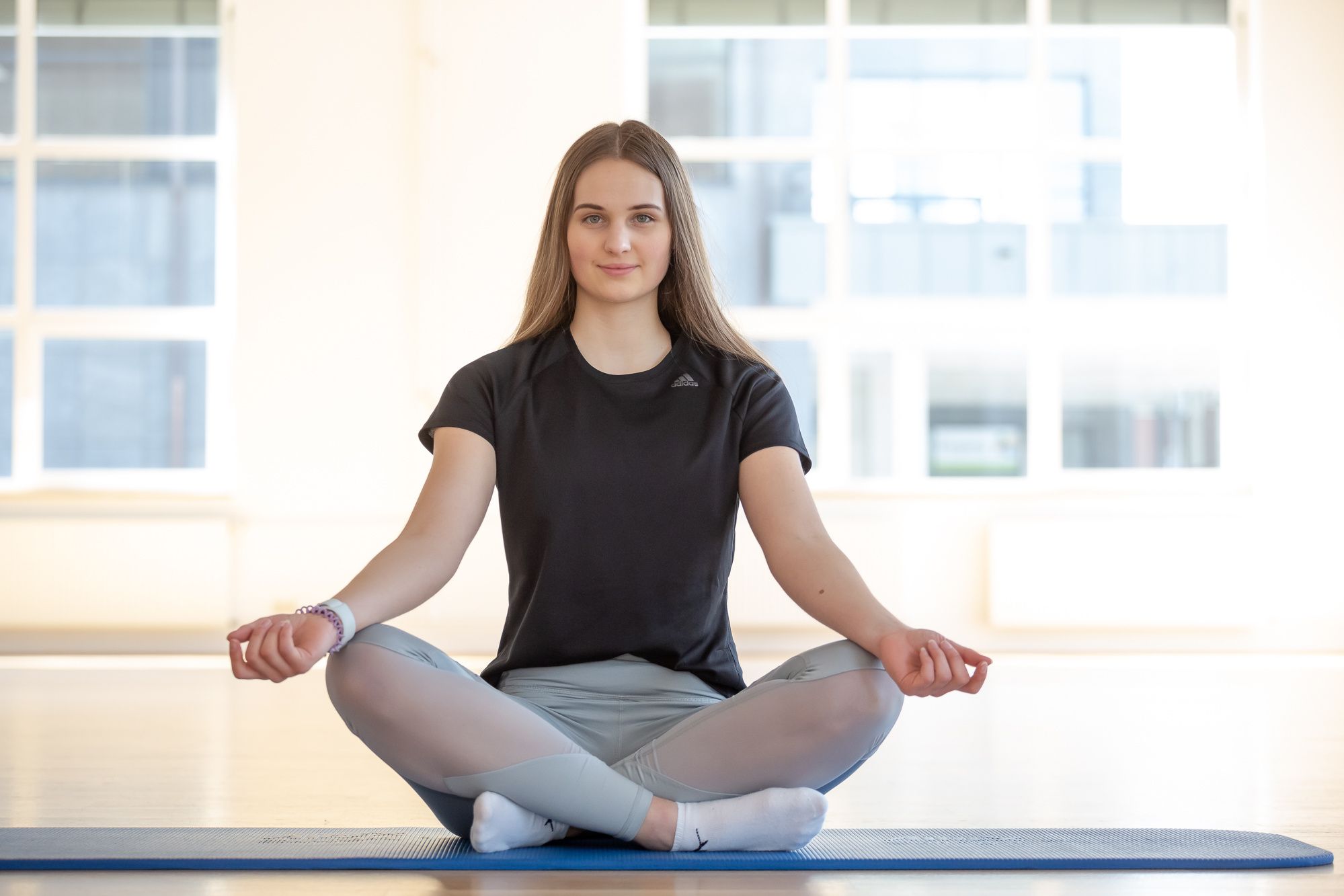 Rabenberg Sportkurse: Yoga In einem hellen Raum sitzt eine junge Frau im Lotussitz auf einer blauen Yogamatte. Sie trägt ein schwarzes T-Shirt und hellgraue Leggings mit weißen Socken. Sie lächelt leicht, und ihre Arme liegen entspannt auf ihren Knien. Im Hintergrund sind helle Fenster zu sehen.