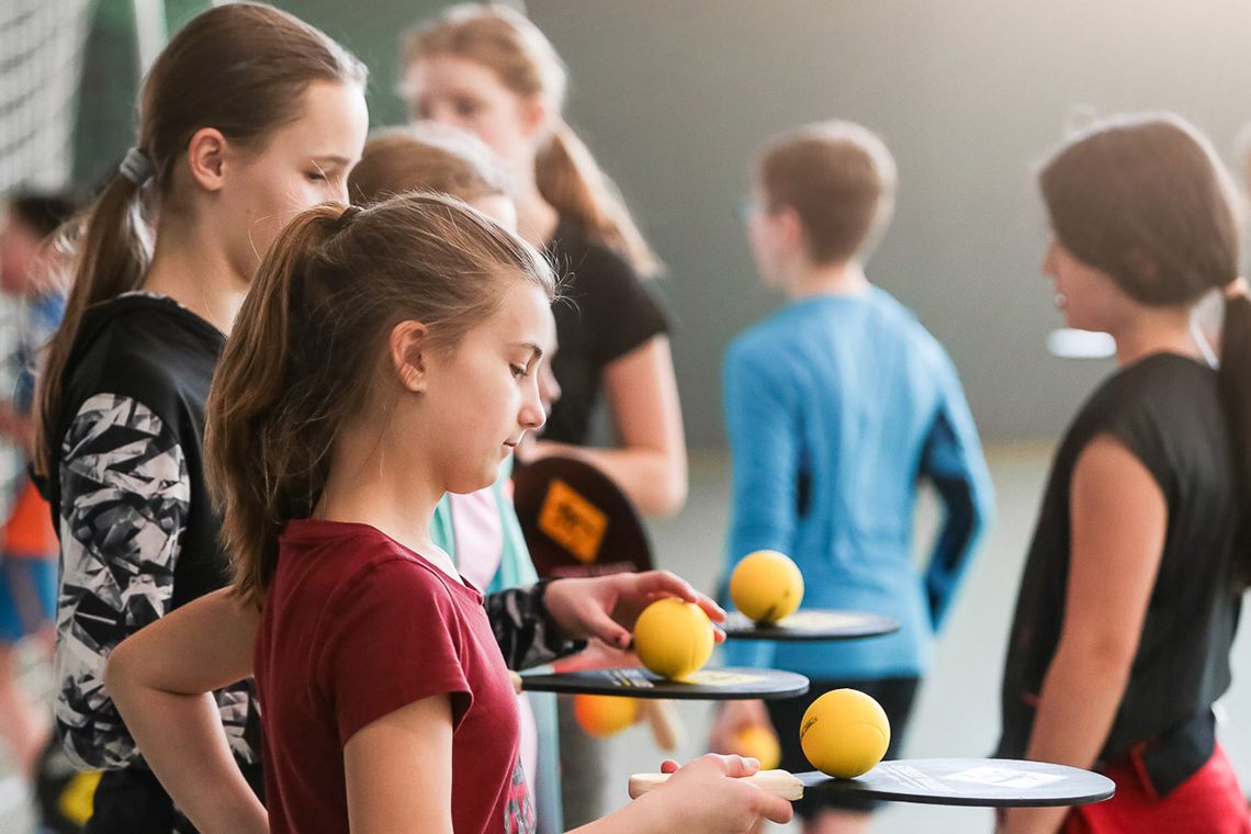 Rabenberg Sportkurse: Trendsportarten In der Aufnahme ist eine Gruppe von Kindern in einer Sporthalle zu sehen, die ein Ballspiel spielen. Etwa sieben Kinder stehen zusammen. Einige von ihnen halten Schläger und gelbe Bälle. Die Mädchen in den Vordergrund halten ihre Schläger und Bälle und bereiten sich darauf vor, zu spielen. Die Halle ist gut beleuchtet.