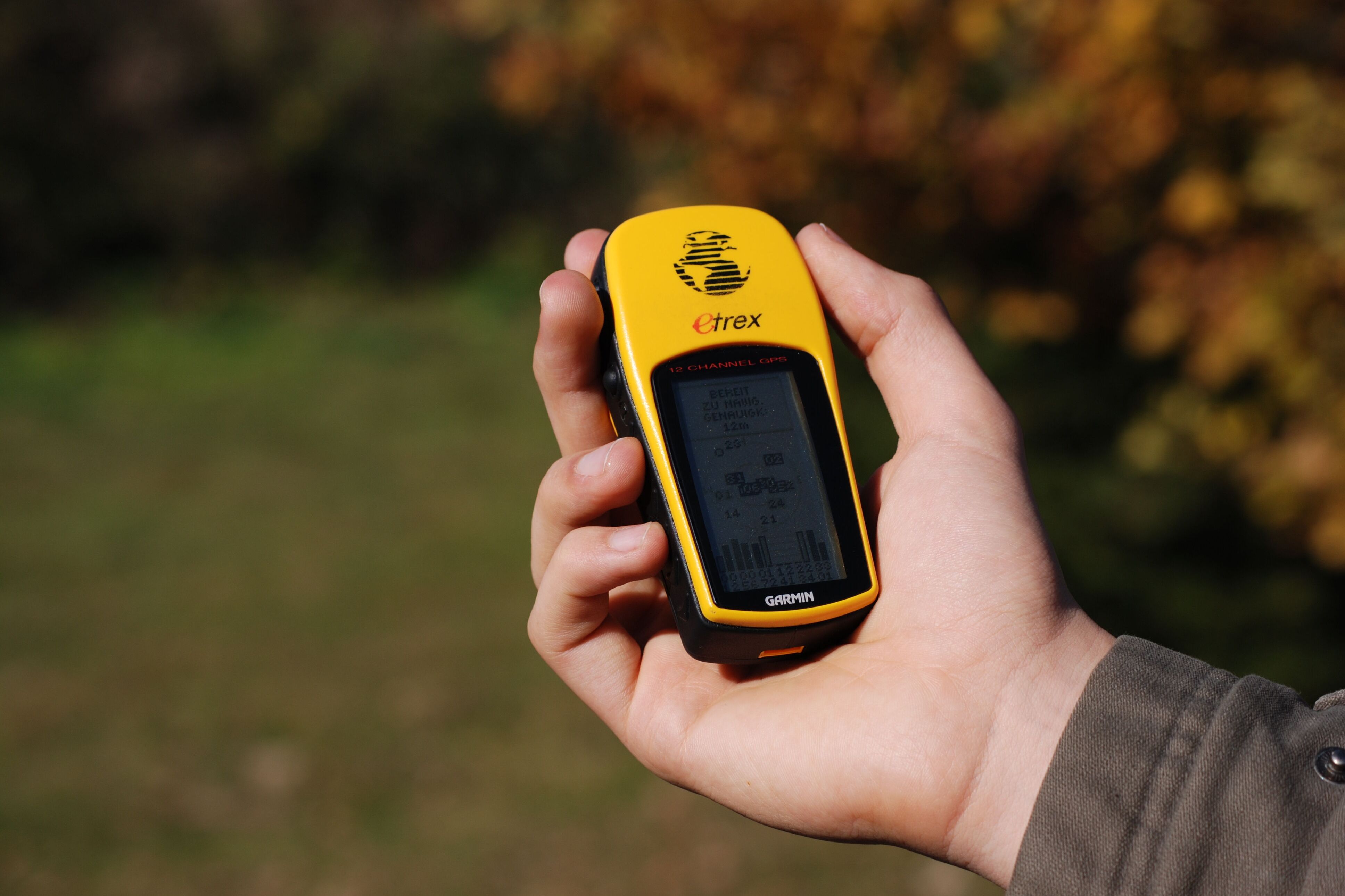 Rabenberg Sportkurse: Naturgeocaching Ein Mann hält ein gelbes GPS-Gerät in seiner Hand. Der Bildschirm des Geräts ist schwarz und zeigt Informationen an. Im Hintergrund ist ein verschwommener Blick auf Bäume mit braunen und grünen Blättern zu sehen. Der Mann trägt ein braunes Hemd.