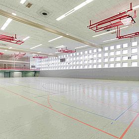 Sportpark Rabenberg: Sporthalle 2 Das Bild zeigt eine Sporthalle. Die Halle ist groß und leer. Der Boden ist hellgrün und mit verschiedenen farbigen Linien markiert, die verschiedene Sportarten anzeigen. Es gibt rote, blaue und schwarze Linien. An der Decke der Halle befinden sich mehrere rote Metallkonstruktionen, die anscheinend zur Aufhängung von Sportgeräten wie Basketballkörben oder Ringen dienen. Die Decke ist weiß und mit rechteckigen Leuchten ausgestattet. An einer Seite der Halle befinden sich eine Reihe von Fenstern, die natürliches Licht hereinlassen. Unterhalb der Fenster befindet sich eine graue Wand. Am Ende der Halle befindet sich eine Anzeigetafel. Auf der linken Seite der Halle befindet sich eine erhöhte Tribüne, die durch ein rotes Geländer geschützt ist. Die Halle wirkt sauber und gut gepflegt. Die Perspektive des Fotos lässt die Halle geräumig und offen erscheinen.