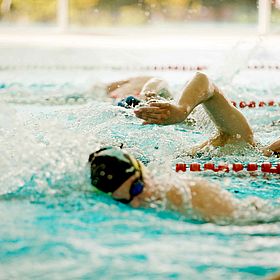 Sportpark Rabenberg: Sommersportwochen Schwimmen Das Bild zeigt eine Schwimmszene. Im Vordergrund sind zwei Schwimmer in einer Schwimmspur zu sehen, die sich im Kraulstil im Wasser bewegen. Die Schwimmer tragen Schwimmbrillen und Badekappen. Einer der Schwimmer hat einen Arm über dem Wasser und den anderen im Wasser. Im Hintergrund sind weitere Schwimmer zu sehen. Das Wasser ist blau und es gibt rote Bojen, die die Schwimmbahnen markieren. Der Hintergrund ist unscharf und zeigt eine helle Decke.
