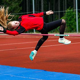 Sportpark Rabenberg: Sommersportwochen Hochsprung Das Bild zeigt ein Mädchen, das Hochsprung macht. Es ist in der Luft und versucht, über die Latte zu springen. Sie trägt eine rote Jacke, schwarze Leggings und Laufschuhe. Ihre Haare sind blond und fliegen in der Luft. Die Latte ist rot und grün. Das Bild wird auf einer roten Sportbahn vor einem grünen Hintergrund aufgenommen. Unter der Latte befindet sich eine blaue Matte.