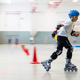 Sportpark Rabenberg: Inlineskating Das Bild zeigt einen Jungen, der auf Inlineskates in einer Halle fährt. Er ist mit einem blauen Helm, Knieschützern, Ellbogenschützern und Handschuhen geschützt. Er trägt ein weißes T-Shirt und eine schwarze Hose. Er fährt mit hoher Geschwindigkeit, was durch die Bewegungsunschärfe im Hintergrund angezeigt wird. Auf dem Boden sind rote Kegel aufgereiht, die wahrscheinlich einen Parcours bilden.