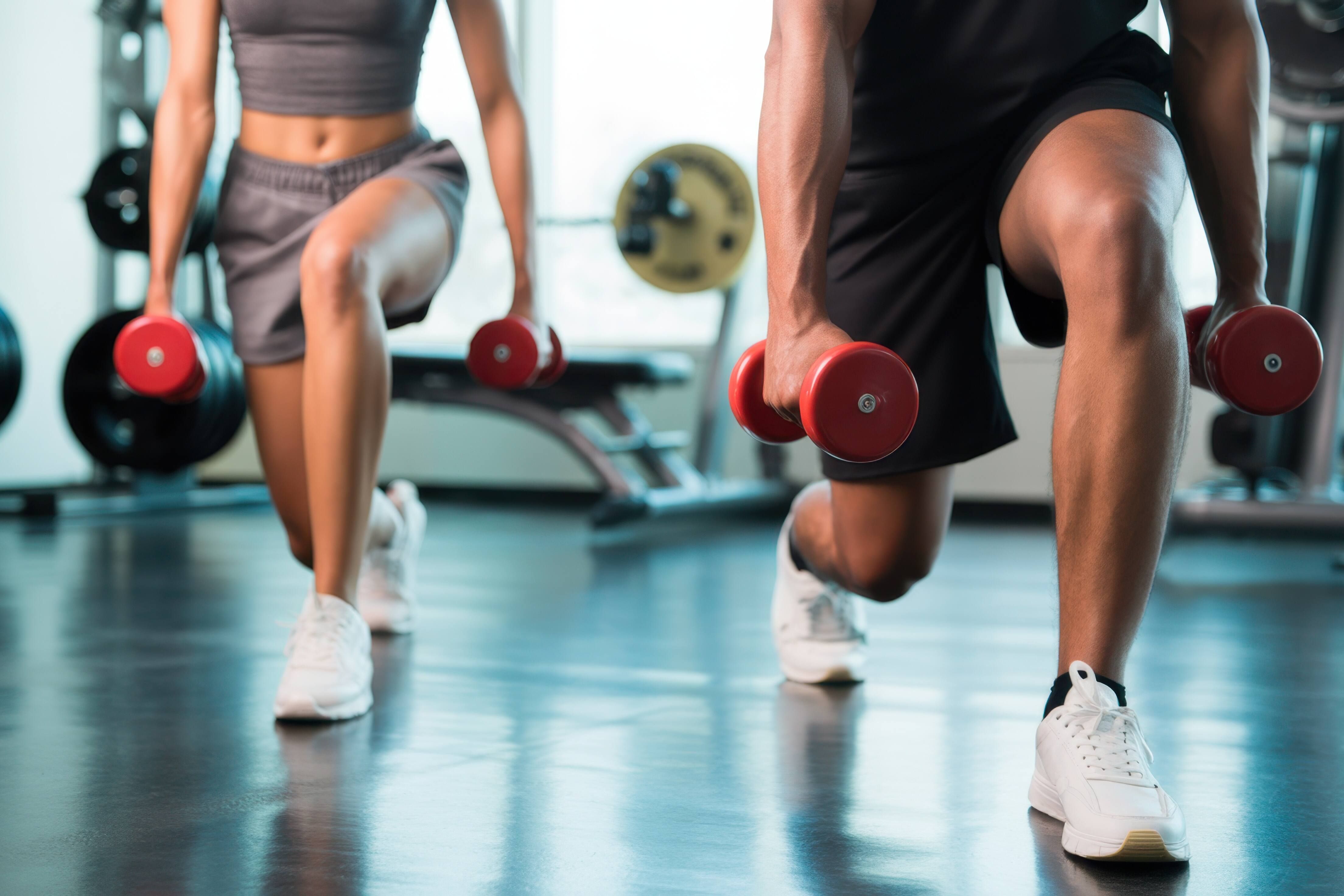 Rabenberg Sportkurse: Lang-/Kurzhantel Das Bild zeigt zwei Personen, die in einem Fitnessstudio Ausfallschritte machen. Beide Personen halten in jeder Hand eine rote Kurzhantel. Die Person im Vordergrund ist ein Mann, dessen Beine und Arme zu sehen sind, er trägt weiße Turnschuhe und schwarze Shorts. Die andere Person im Hintergrund ist eine Frau, deren Oberkörper und Beine zu sehen sind, sie trägt graue Shorts und ein graues Top und weiße Turnschuhe. Im Hintergrund sind Fitnessgeräte zu sehen.