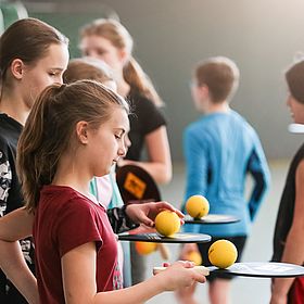 Sportpark Rabenberg: Sommersportwochen Sportspiele Das Bild zeigt eine Gruppe von Kindern in einer Sporthalle. Sie scheinen gerade eine Sportart zu spielen, bei der sie kleine gelbe Bälle mit Schlägern schlagen. Im Vordergrund stehen mehrere Mädchen die jeweils einen Ball auf dem Schläger balancieren. Eines der Mädchen in einem roten T-Shirt hält einen Schläger in der Hand und legt gerade einen gelben Ball auf den Schläger. Andere Mädchen halten ebenfalls Schläger und Bälle. Im Hintergrund sind weitere Kinder zu sehen, die in verschiedene Richtungen schauen oder sich bewegen.