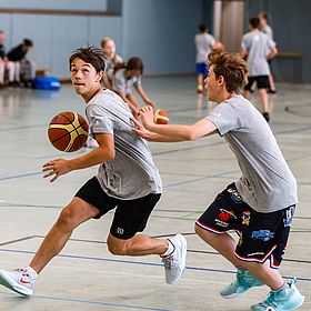 Sportpark Rabenberg: Sommersportwochen Basketball Das Bild zeigt zwei Jungen, die in einer Turnhalle Basketball spielen. Der Junge im Vordergrund dribbelt den Ball mit der rechten Hand und blickt nach links. Er trägt ein graues T-Shirt, schwarze Shorts und weiße Turnschuhe mit roten Akzenten. Der Ball ist orange-braun mit schwarzen Rillen. Der andere Junge versucht, ihm den Ball abzunehmen und streckt seine Hand nach dem ersten Jungen aus. Er trägt ein graues T-Shirt, Basketballshorts mit Teamlogos und hellblaue Turnschuhe. Im Hintergrund sind weitere Personen zu sehen, einige davon scheinen ebenfalls Basketball zu spielen, während andere zuschauen. Sie tragen ähnliche Outfits wie die beiden Jungen im Vordergrund. Auf dem Boden sind Linien für das Spielfeld aufgezeichnet.