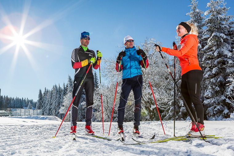 Rabenberg Sportkurse: Ski-Unterricht Das Foto zeigt drei Personen auf Langlaufskiern in einer verschneiten Winterlandschaft. Im Hintergrund leuchten Bäume, der Himmel ist blau. Die Sonne scheint hell und wirft Sonnenstrahlen. Die drei Skifahrer sind in Sportkleidung gekleidet und scheinen sich zu unterhalten oder eine Pause zu machen.