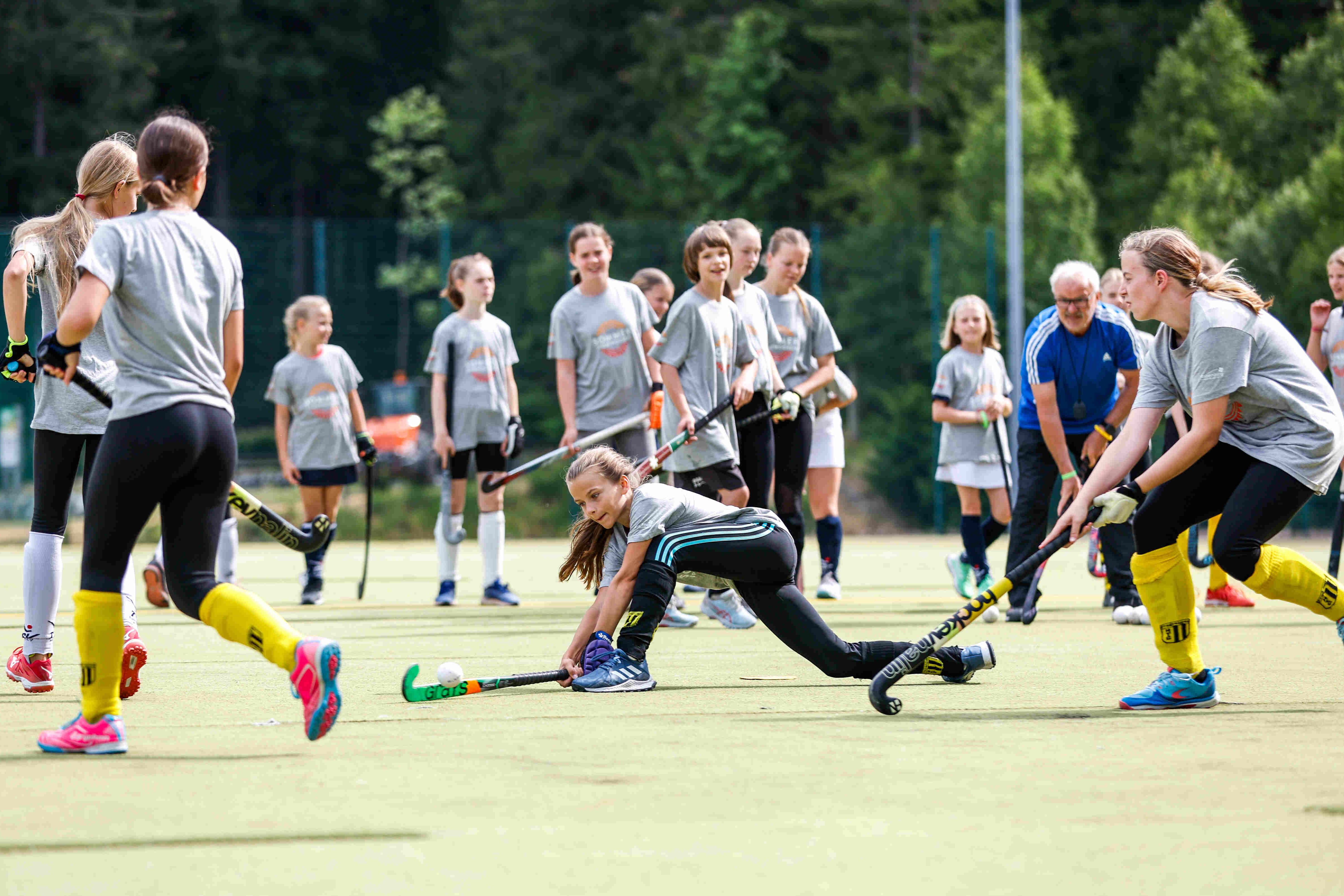 Sportpark Rabenberg: Sommersportwochen Feldhockey Das Bild zeigt eine Gruppe junger Mädchen, die Hockey spielen. Sie tragen alle graue T-Shirts, schwarze Leggings und farbige Socken und Schuhe. Einige Mädchen tragen gelbe Stutzen. Einige Mädchen haben ihre Haare zu einem Pferdeschwanz zusammengebunden. Auf dem grünen Rasen stehen auch einige Mädchen und beobachten das Spiel. Im Hintergrund stehen Bäume und ein älterer Mann in einem blauen T-Shirt. Das Wetter ist sonnig. Ein Mädchen ist in einer niedrigen Position abgebildet, während es versucht, den Hockeyball mit seinem Schläger zu treffen. Die anderen Mädchen stehen im Hintergrund und beobachten das Spiel. Der Mann im blauen T-Shirt scheint ein Trainer zu sein.