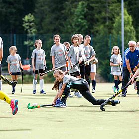 Sportpark Rabenberg: Sommersportwochen Feldhockey Das Bild zeigt eine Gruppe junger Mädchen, die Hockey spielen. Sie tragen alle graue T-Shirts, schwarze Leggings und farbige Socken und Schuhe. Einige Mädchen tragen gelbe Stutzen. Einige Mädchen haben ihre Haare zu einem Pferdeschwanz zusammengebunden. Auf dem grünen Rasen stehen auch einige Mädchen und beobachten das Spiel. Im Hintergrund stehen Bäume und ein älterer Mann in einem blauen T-Shirt. Das Wetter ist sonnig. Ein Mädchen ist in einer niedrigen Position abgebildet, während es versucht, den Hockeyball mit seinem Schläger zu treffen. Die anderen Mädchen stehen im Hintergrund und beobachten das Spiel. Der Mann im blauen T-Shirt scheint ein Trainer zu sein.