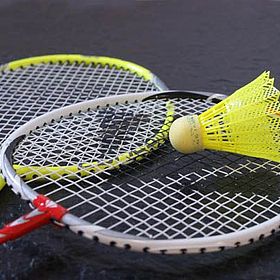 Sportpark Rabenberg: Sommersportwochen Badminton Das Bild zeigt zwei Badmintonschläger und einen gelben Federball. Die Schläger sind in einer diagonalen Anordnung angeordnet, wobei sich der vordere Schläger über dem hinteren befindet. Der Griff des vorderen Schlägers ist rot und weiß, während der Griff des hinteren Schlägers gelb ist. Die Rahmen der Schläger sind silberfarben. Das Netz der Schläger ist weiß. Der Federball liegt auf dem Netz des vorderen Schlägers. Der Federball hat einen gelben Federkopf und einen hellgelben Korkfuß. Der Hintergrund des Bildes ist dunkel.