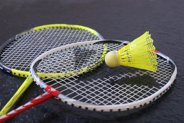 Sportpark Rabenberg: Sommersportwochen Badminton Das Bild zeigt zwei Badmintonschläger und einen gelben Federball. Die Schläger sind in einer diagonalen Anordnung angeordnet, wobei sich der vordere Schläger über dem hinteren befindet. Der Griff des vorderen Schlägers ist rot und weiß, während der Griff des hinteren Schlägers gelb ist. Die Rahmen der Schläger sind silberfarben. Das Netz der Schläger ist weiß. Der Federball liegt auf dem Netz des vorderen Schlägers. Der Federball hat einen gelben Federkopf und einen hellgelben Korkfuß. Der Hintergrund des Bildes ist dunkel.
