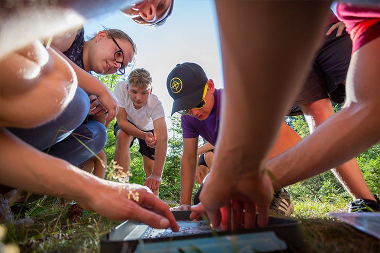 Rabenberg Sportkurse: Teamspiele Das Bild zeigt eine Gruppe von Menschen, die sich um etwas auf dem Boden versammelt haben. Aus der Perspektive des Betrachters blickt man von unten nach oben in die Szene hinein. Im Zentrum des Geschehens liegt vermutlich eine flache Schale oder ein Tablett, an dem mehrere Personen arbeiten. Die Personen sind jung und tragen Freizeitkleidung. Einige haben die Köpfe gesenkt, um sich die Arbeit anzusehen. Eine Person trägt eine Sonnenbrille und eine Baseballkappe. Im Hintergrund ist etwas Grün zu sehen, vermutlich Gras oder Bäume, und der Himmel ist blau.