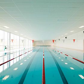 Sportpark Rabenberg: Schwimmhalle 50 Meter Das Bild zeigt ein leeres Schwimmbecken in einem Innenraum. Das Schwimmbecken ist von einer rechteckigen Form und hat klares, türkisfarbenes Wasser. Auf beiden Seiten des Beckens verlaufen rote und weiße Schwimmbahnmarkierungen. Die Wände des Raumes sind weiß und mit quadratischen Fliesen bedeckt. An der Decke sind rechteckige, eingebaute Leuchten angebracht. Große Fenster erstrecken sich entlang einer Seite des Raumes und lassen Tageslicht herein. Am Ende des Beckens befinden sich einige rote und weiße Flaggen, die an Schnüren hängen. Insgesamt ist das Bild sauber und gut beleuchtet, was einen Eindruck von einem modernen und gepflegten Schwimmbad vermittelt.