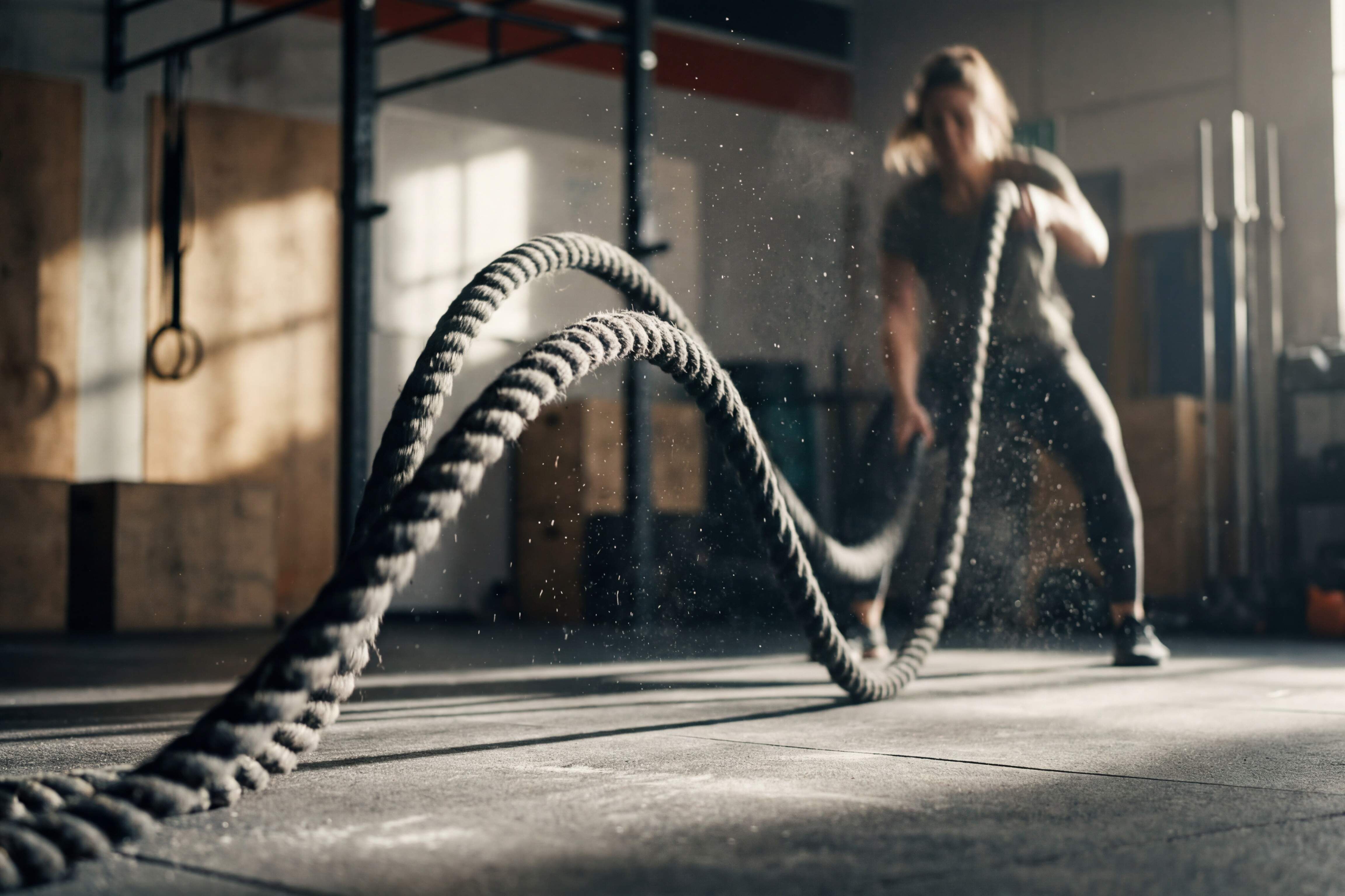 Rabenberg Sportkurse: Hochintensives Intervalltraining Das Bild zeigt eine unscharfe Person in einem Fitnessstudio, die trainiert. Sie hält zwei Seile in den Händen, eins in jeder Hand und scheint diese im Wechsel schnell auf und ab zu bewegen, sodass Staubpartikel durch die Luft wirbeln. Der Hintergrund ist unscharf, aber es ist möglich, Geräte wie Gewichte und ein Fenster zu sehen.