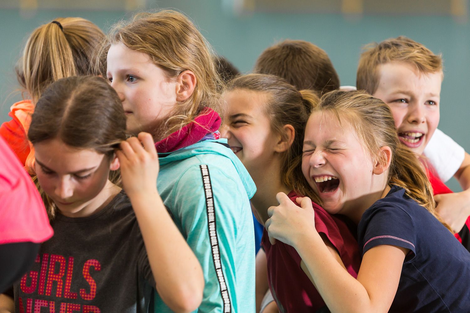 Sportpark Rabenberg: Sommersportwochen Freizeitprogramm Das Bild zeigt eine Gruppe von Kindern, die scheinbar viel Spaß haben. Sie stehen eng beieinander und einige von ihnen lachen aus vollem Halse. Im Vordergrund ist ein Mädchen mit dunklem Haar zu sehen. Daneben stehen weitere Kinder, darunter ein Mädchen mit hellblondem Haar und einem türkisfarbenen Hoodie. Direkt daneben lacht ein Mädchen, das von einem anderen Mädchen umarmt wird. Der Junge rechts im Bild lacht ebenfalls breit.