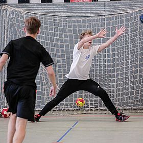 Sportpark Rabenberg: Sommersportwochen Handball Das Bild zeigt eine Szene aus einem Handballspiel. Im Vordergrund sind zwei Jungen zu sehen. Einer der Jungen steht mit dem Rücken zur Kamera und trägt ein schwarzes T-Shirt und eine schwarze Hose. Er scheint gerade einen Wurf auszuführen. Der andere Junge steht im Tor und versucht, den Ball zu fangen. Er trägt ein weißes T-Shirt und eine schwarze Hose. Er hat die Arme weit geöffnet und versucht, den Ball abzuwehren. Der Ball ist gerade im Flug und nähert sich dem Tor. Im Hintergrund ist ein Handballtor zu sehen, das aus einem Netz und einem Rahmen besteht. Auf dem Boden liegen weitere Bälle. Die Halle hat graue Wände.