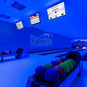 Sportpark Rabenberg: Schwarzlicht Bowlingbahn Das Bild zeigt eine Bowlingbahn in bläulichem Licht. Im Vordergrund befinden sich zwei Kugellager mit farbigen Bowlingkugeln. Am Ende der Bahnen stehen die Kegel, dahinter ist eine Wandmalerei mit Berglandschaft und Skifahrern zu erkennen. Über den Bahnen sind mehrere Monitore an der Decke angebracht, die Informationen über das Bowling anzeigen. Neben der linken Bowlingbahn ist das Logo „Rabenberg“ an der Wand angebracht.