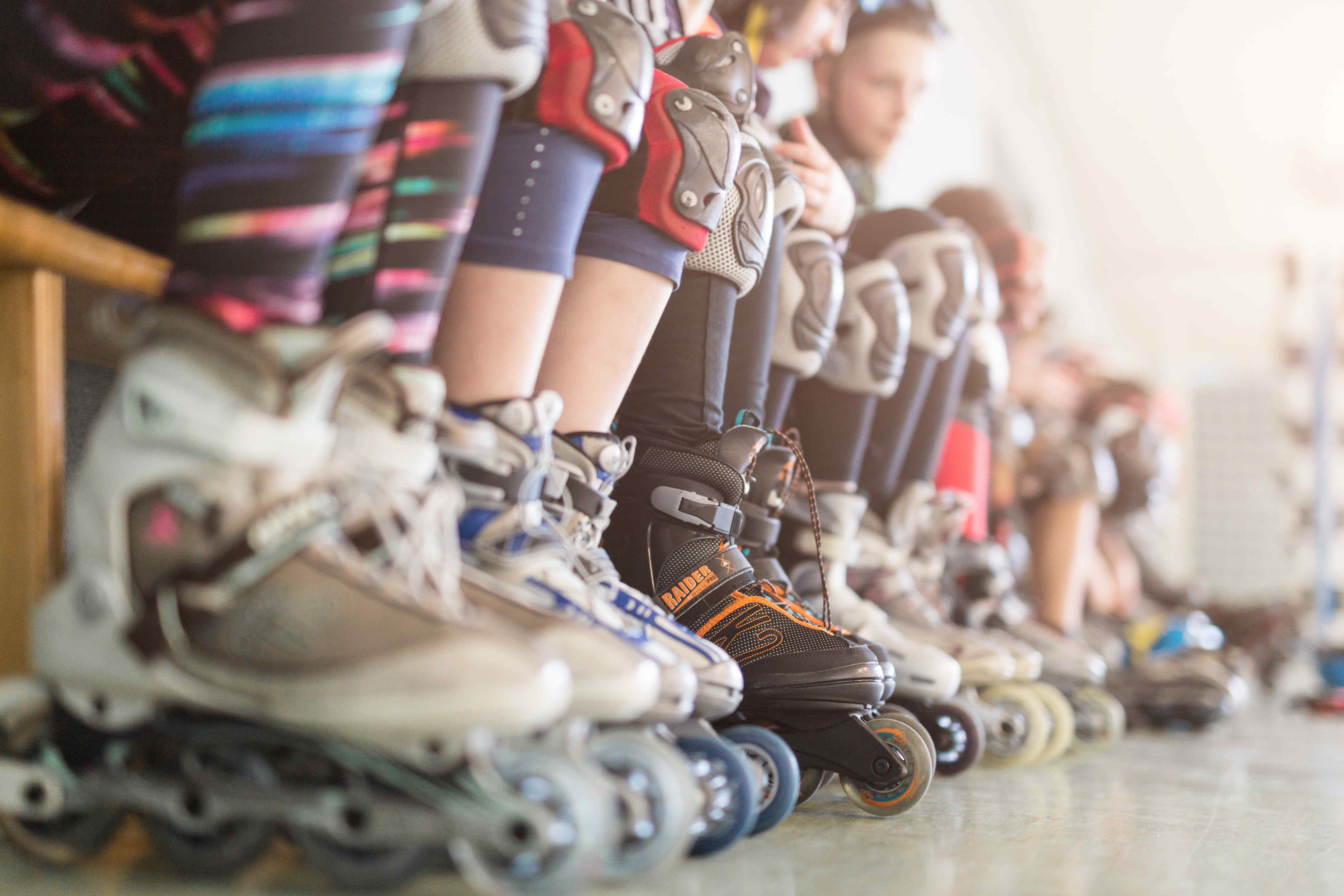 Rabenberg Sportkurse: Inlineskating Das Bild zeigt eine Reihe von Inline-Skates in einer Reihe. Die Skates sind in verschiedenen Farben gehalten und stehen auf einer hellen Oberfläche. Die Füße der Skater sind teilweise zu sehen, wobei Schutzkleidung wie Knieschützer und Strumpfhosen getragen werden. Der Hintergrund ist unscharf, was den Fokus auf die Inline-Skates lenkt.