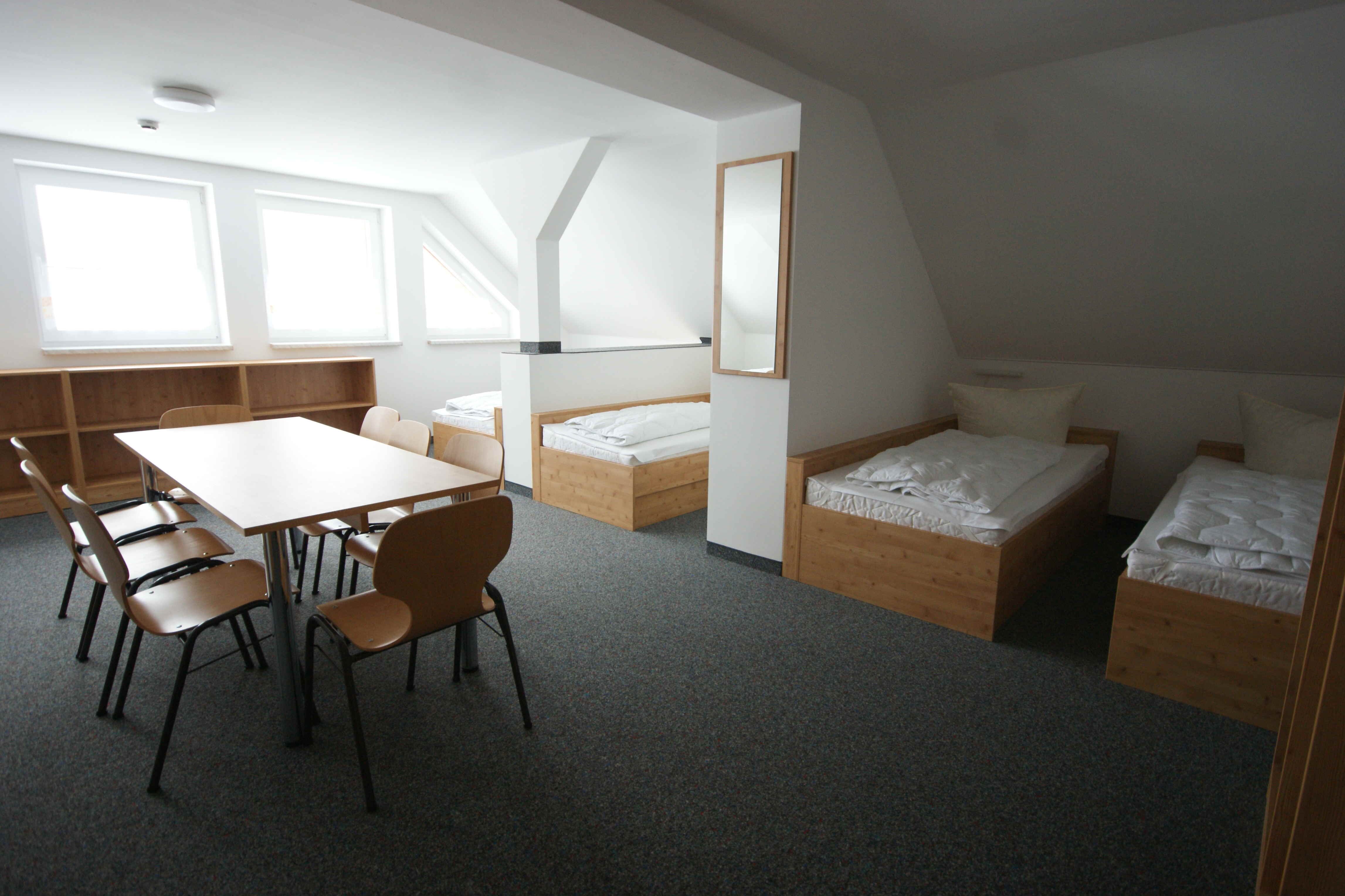 Sportpark Rabenberg: Mehrbettzimmer Haus 8 Das Bild zeigt einen Innenraum, der wie ein Schlafsaal oder ein Gästezimmer aussieht. Der Raum ist hell und mit neutralen Farben gehalten. Im Vordergrund befindet sich ein Tisch mit sechs Stühlen. Der Tisch ist rechteckig und hat eine helle, glatte Oberfläche. Die Stühle sind aus Holz und haben schwarze Metallbeine. Im Hintergrund befinden sich mehrere Betten. Die Betten sind aus Holz und haben weiße Bettwäsche. Es gibt auch einen Spiegel an der Wand. Der Boden ist mit einem grauen Teppich ausgelegt. Die Wände sind weiß.
