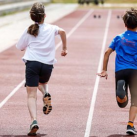 Sportpark Rabenberg: Sommersportwochen Sprint Das Bild zeigt zwei Kinder, die auf einer roten Laufbahn rennen. Beide sind von hinten zu sehen. Das Mädchen, das links auf dem Bild rennt, trägt ein weißes T-Shirt und schwarze Shorts. Sie hat ihr Haar zu einem Pferdeschwanz zusammengebunden. Ihre Arme sind angewinkelt. Der Junge, der rechts auf dem Bild rennt, trägt ein blaues T-Shirt mit einem Logo auf der Rückseite. Er trägt schwarze Shorts und schwarze Schuhe mit orangefarbenen Socken. Seine Arme sind angewinkelt. Die Laufbahn hat weiße Markierungen.