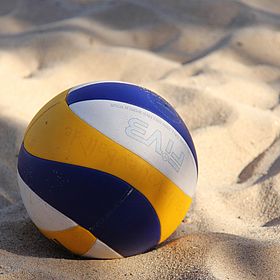 Sportpark Rabenberg: Beachvolleyball im Sand Das Bild zeigt einen Volleyball, der im Sand liegt. Der Ball ist eine Kombination aus Blau, Weiß und Gelb. Das Logo "FIVB" ist auf dem Ball zu sehen. Der Sand ist hell und weist eine unebene Textur auf.