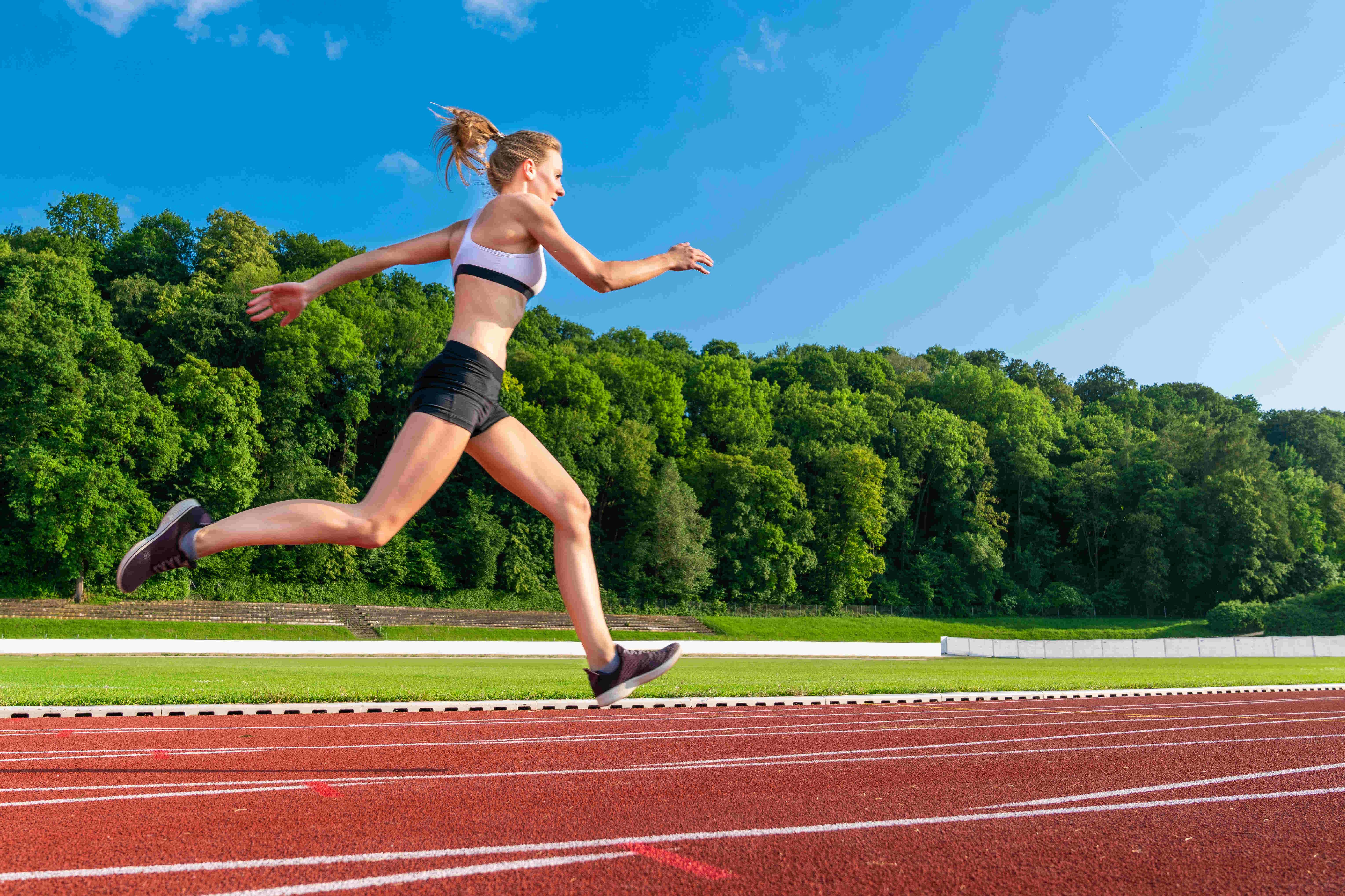 Rabenberg Sportkurse: Athletik Training Das Bild zeigt eine Frau, die auf einer roten Laufbahn rennt. Sie ist in Bewegung abgebildet, mit einem Bein in der Luft und dem anderen, das den Boden berührt. Sie trägt Sportkleidung, darunter einen weißen Sport-BH und schwarze Shorts. Im Hintergrund befinden sich eine grüne Wiese und Bäume unter einem blauen Himmel.