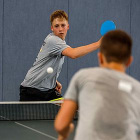 Sportpark Rabenberg: Sommersportwochen Tischtennis Das Foto zeigt zwei Jungen, die Tischtennis spielen. Der Junge im Vordergrund ist unscharf und von hinten zu sehen. Er trägt ein graues T-Shirt. Der andere Junge, der im Fokus steht, ist auch mit einem grauen T-Shirt bekleidet und schaut in Richtung der Kamera. Er hat einen blonden Haarschnitt. Er hält einen blauen Schläger in der Hand und schlägt gerade den Ball. Der Ball ist auch im Fokus und befindet sich in der Mitte des Bildes. Im Hintergrund befindet sich eine blaue Wand und ein Tischtennisnetz.