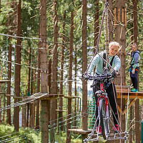 Sportpark Rabenberg: Abenteuerpark Fahrrad Das Bild zeigt eine junge Frau, die mit einem Fahrrad über einen Hochseilgarten fährt. Sie ist in einer grünen Jacke und schwarzen Hosen gekleidet und trägt Handschuhe. Sie hat ein breites Lächeln im Gesicht und schaut in die Kamera. Das Fahrrad ist rot und hat dicke Reifen. Im Hintergrund ist ein anderer junger Mensch zu sehen, der auf einer Plattform steht und ebenfalls einen Gurt trägt. Beide sind von Bäumen umgeben, und es gibt eine Reihe von Hindernissen, die durch den Hochseilgarten führen.