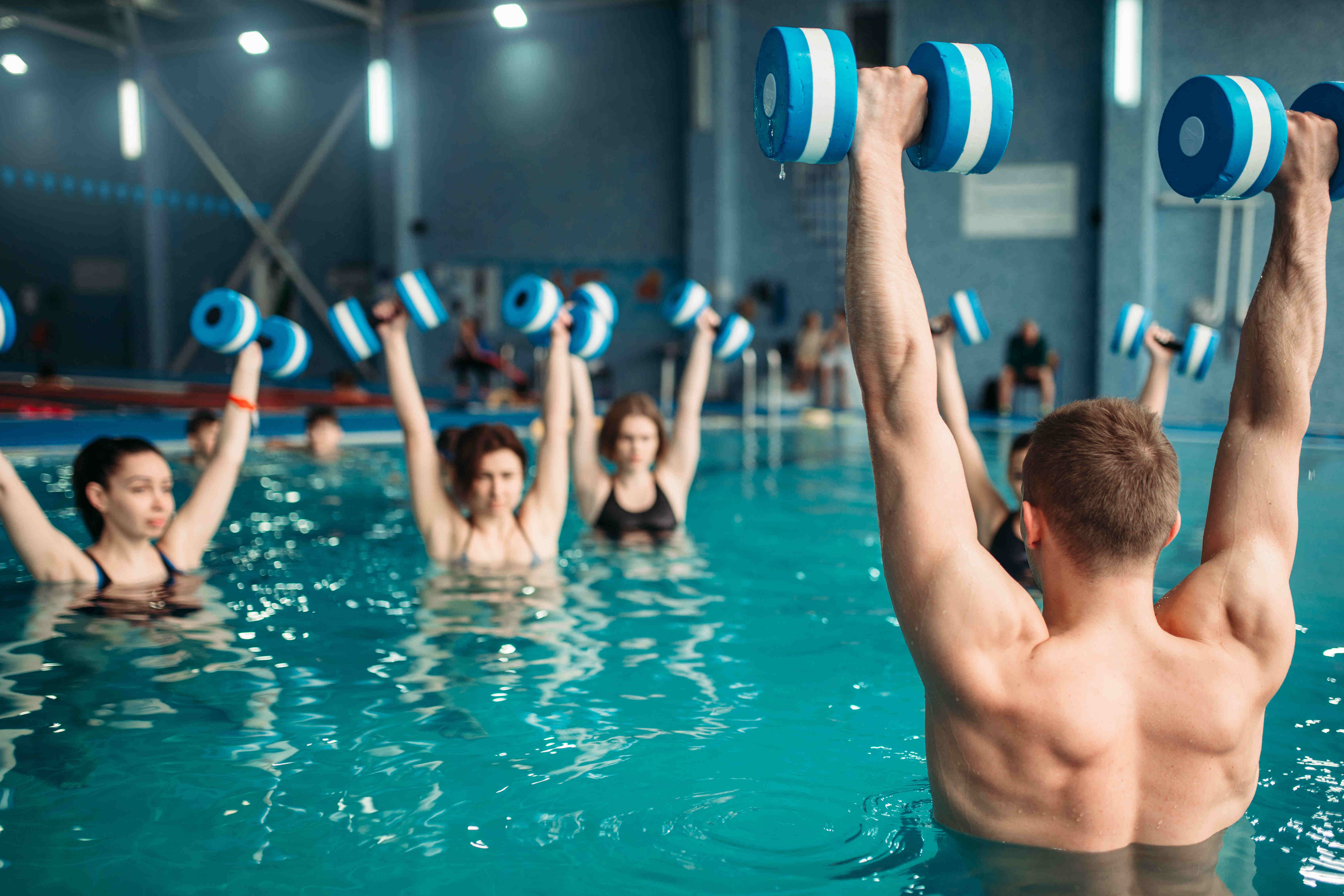 Rabenberg Sportkurse: Aqua-Cross Das Bild zeigt eine Gruppe von Menschen, die im Wasser trainieren. Sie halten blaue Hanteln hoch in die Luft. Die Personen sind im Wasser bis zur Brust. Der Hintergrund ist eine Schwimmhalle mit blauen Wänden und einigen Zuschauern.