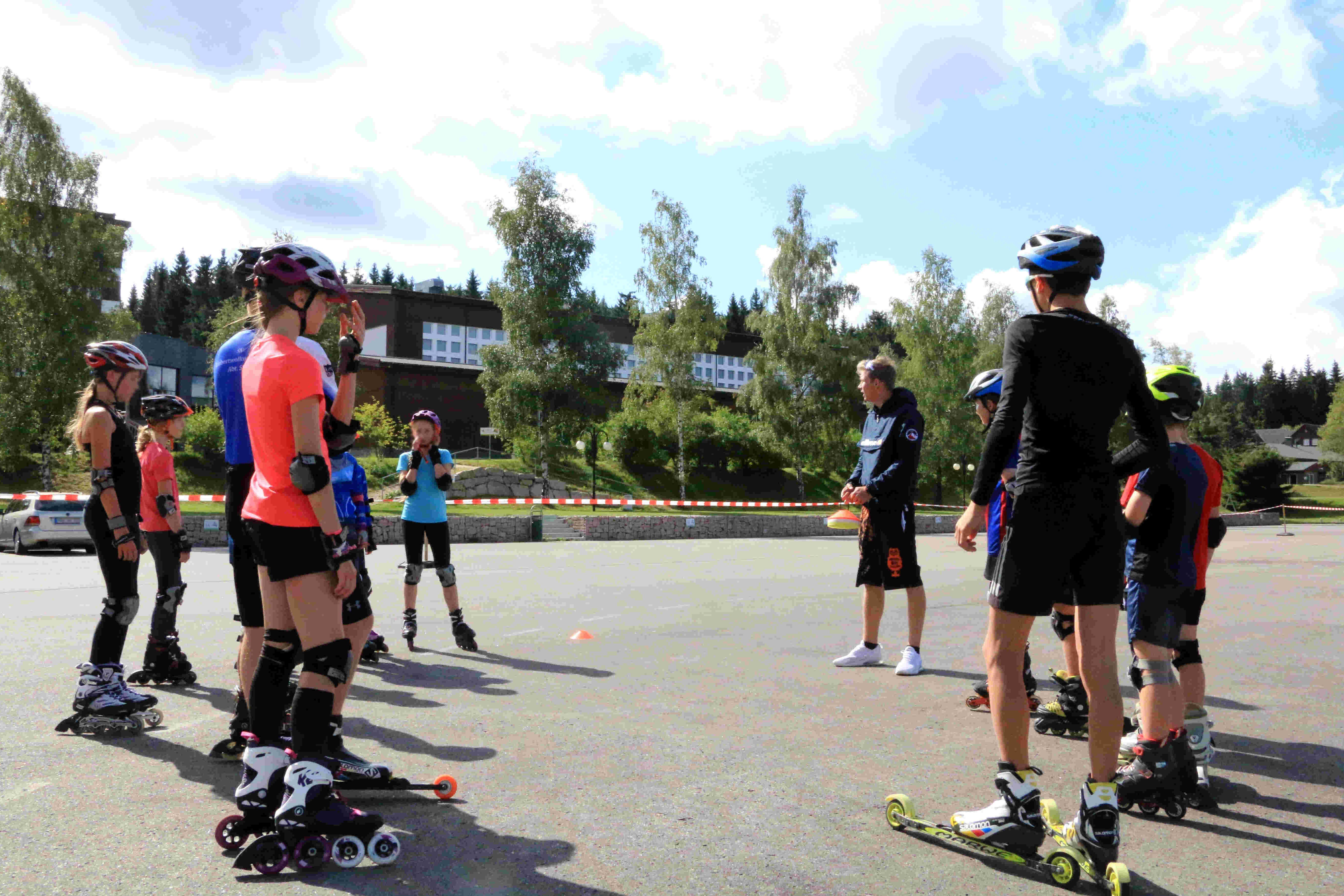 Sportpark Rabenberg: Sommersportwochen Sommerski Das Bild zeigt eine Gruppe von Menschen, die auf einer asphaltierten Fläche Inlineskaten oder Rollski fahren. Die Gruppe besteht aus Kindern und Jugendlichen, sowie einem Mann. Die meisten tragen Helme, Knieschützer und Ellbogenschützer, was darauf hindeutet, dass sie sich auf Sicherheit konzentrieren. Im Hintergrund sind Bäume und Gebäude zu sehen, was darauf hindeutet, dass die Szene im Freien stattfindet. Der Himmel ist blau und sonnig. Die Leute scheinen sich in einem Training oder einer Unterrichtsstunde zu befinden, da ein Mann in der Mitte der Gruppe steht und Anweisungen zu geben scheint.