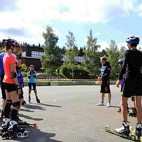 Sportpark Rabenberg: Sommersportwochen Sommerski Das Bild zeigt eine Gruppe von Menschen, die auf einer asphaltierten Fläche Inlineskaten oder Rollski fahren. Die Gruppe besteht aus Kindern und Jugendlichen, sowie einem Mann. Die meisten tragen Helme, Knieschützer und Ellbogenschützer, was darauf hindeutet, dass sie sich auf Sicherheit konzentrieren. Im Hintergrund sind Bäume und Gebäude zu sehen, was darauf hindeutet, dass die Szene im Freien stattfindet. Der Himmel ist blau und sonnig. Die Leute scheinen sich in einem Training oder einer Unterrichtsstunde zu befinden, da ein Mann in der Mitte der Gruppe steht und Anweisungen zu geben scheint.