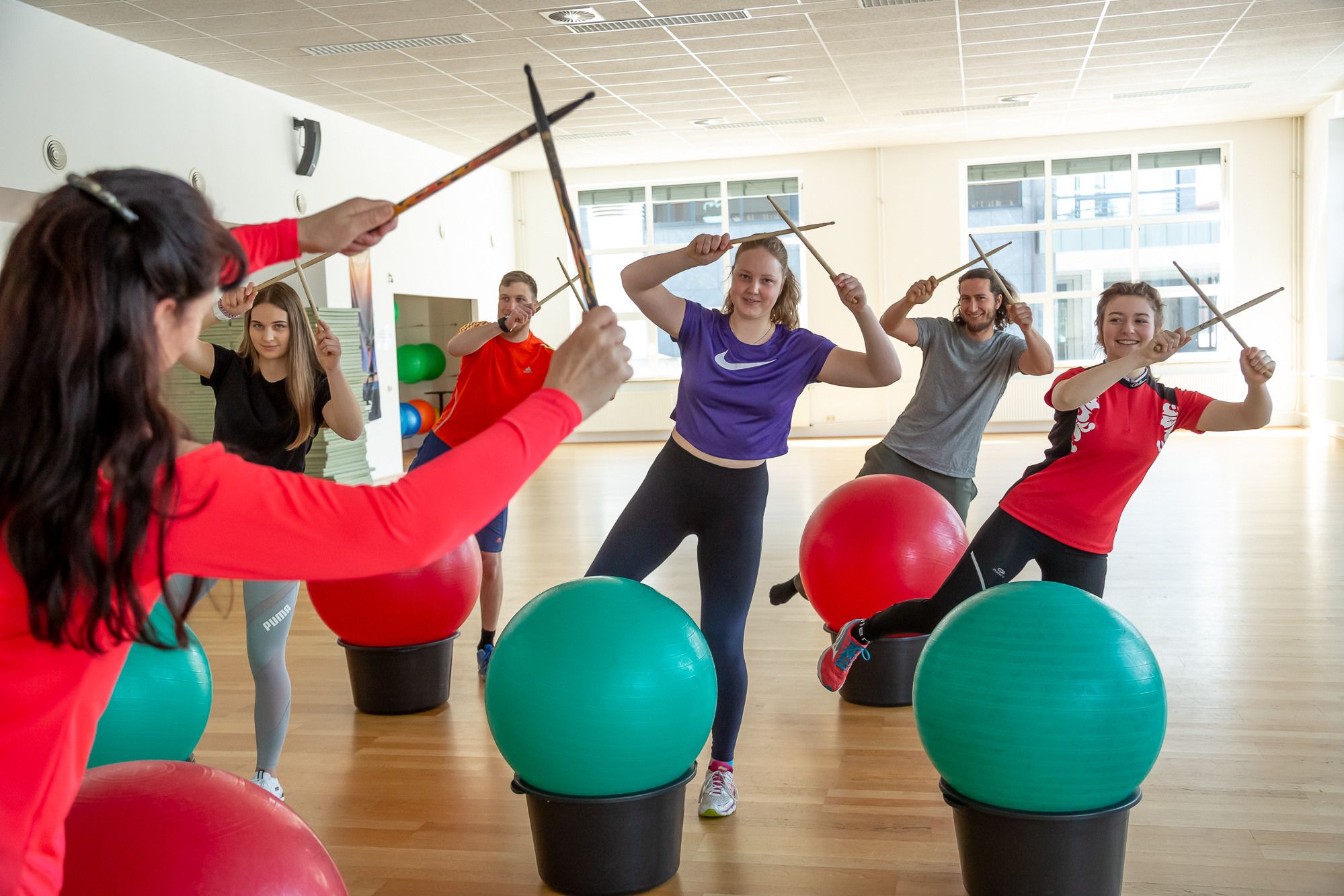 Rabenberg Sportkurse: Drums Alive® Das Bild zeigt eine Gruppe von sechs Personen in einem Fitnessstudio. Alle halten Drumsticks in den Händen und stehen anscheinend über Gymnastikbällen. Die Personen sind in Sportkleidung gekleidet und scheinen in Bewegung zu sein. Im Hintergrund sind Fenster und weitere Gymnastikbälle zu sehen. Die Atmosphäre wirkt lebendig und sportlich.