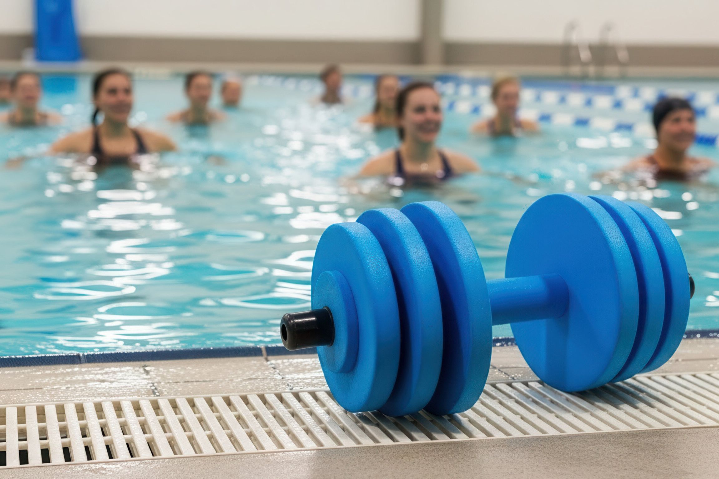 Rabenberg Sportkurse: Aqua-Workout Das Bild zeigt eine Gruppe von Menschen, die Wassergymnastik machen. Im Vordergrund befindet sich eine blaue Hantel. Im Hintergrund befinden sich die Personen im Wasser.