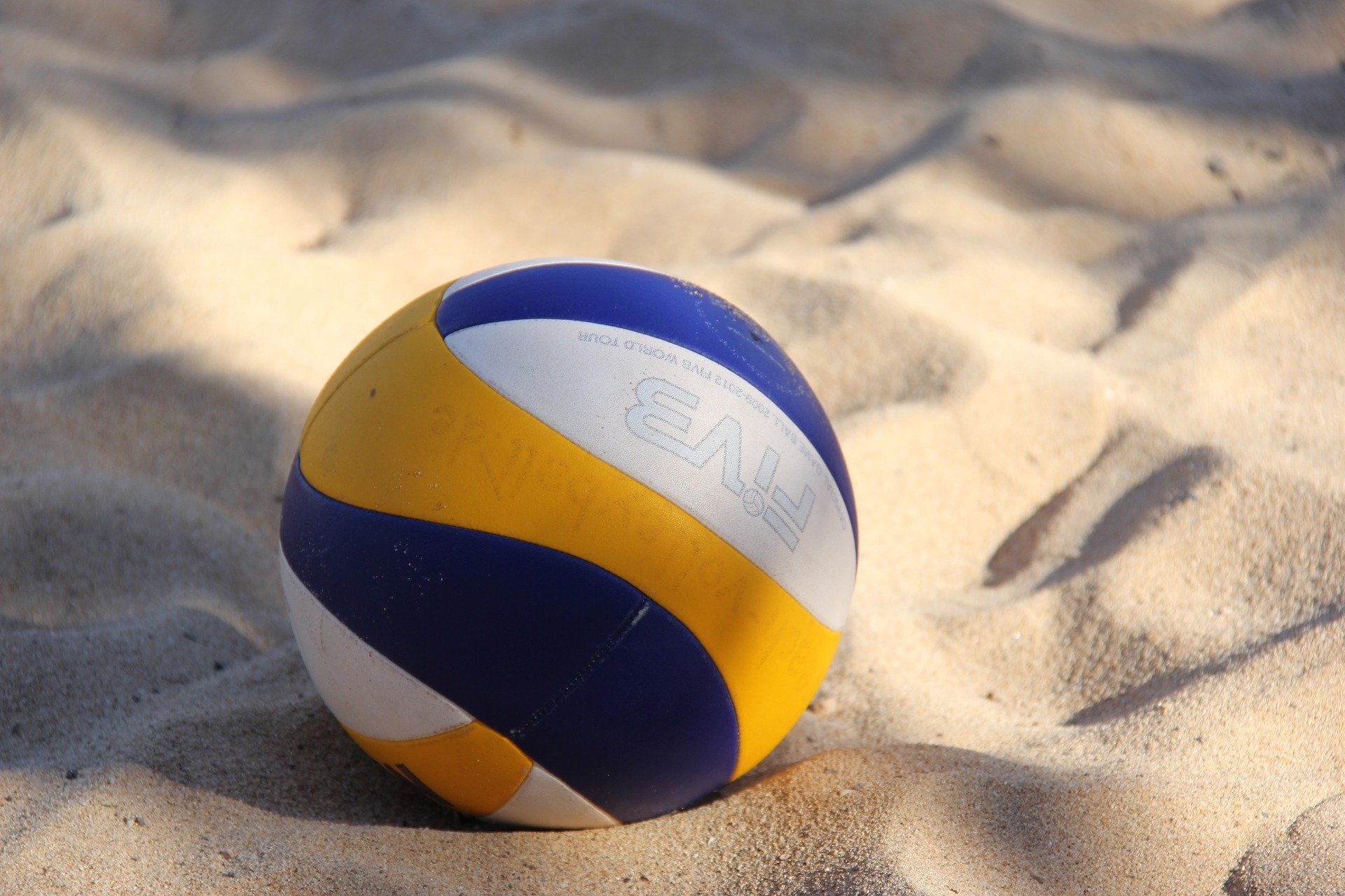 Sportpark Rabenberg: Beachvolleyball im Sand Das Bild zeigt einen Volleyball, der im Sand liegt. Der Ball ist eine Kombination aus Blau, Weiß und Gelb. Das Logo "FIVB" ist auf dem Ball zu sehen. Der Sand ist hell und weist eine unebene Textur auf.
