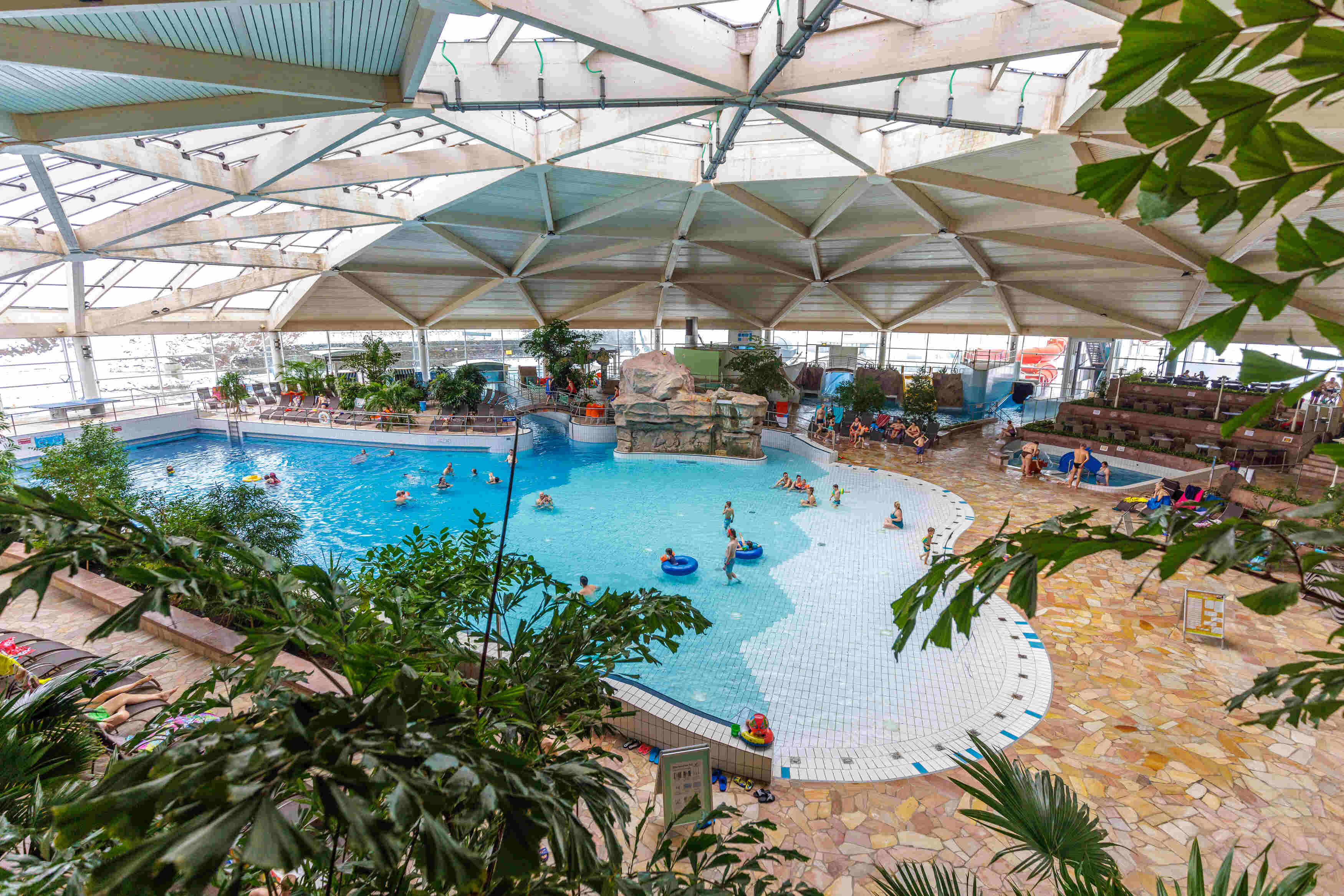 Freizeitbad an den Greifensteinen Geyer Innenaufnahme ©Ronny Küttner/photoron Das Bild zeigt eine Indoor-Badelandschaft. Im Vordergrund sind grüne Blätter zu sehen, die einen Teil der Szene verbergen. Im Mittelpunkt des Bildes befindet sich ein großes Schwimmbecken mit blauem Wasser. Es gibt mehrere Personen im Becken, die schwimmen oder sich entspannen. Einige von ihnen benutzen Schwimmreifen. Das Becken hat eine unregelmäßige Form und ist von einem weißen Boden mit blauen Fliesen umgeben. Um das Schwimmbecken herum befinden sich verschiedene Bereiche. Auf der rechten Seite gibt es einen Bereich mit Liegestühlen, auf denen sich einige Leute entspannen. Es gibt auch einen Bereich mit einem Felsen und einer Brücke, der wie ein Wasserfall aussieht. Weiter rechts ist eine Rutsche zu sehen. Die Decke des Schwimmbades ist aus Glas und Holz gebaut und hat eine interessante geometrische Form. Durch die Decke fällt Tageslicht in den Raum.