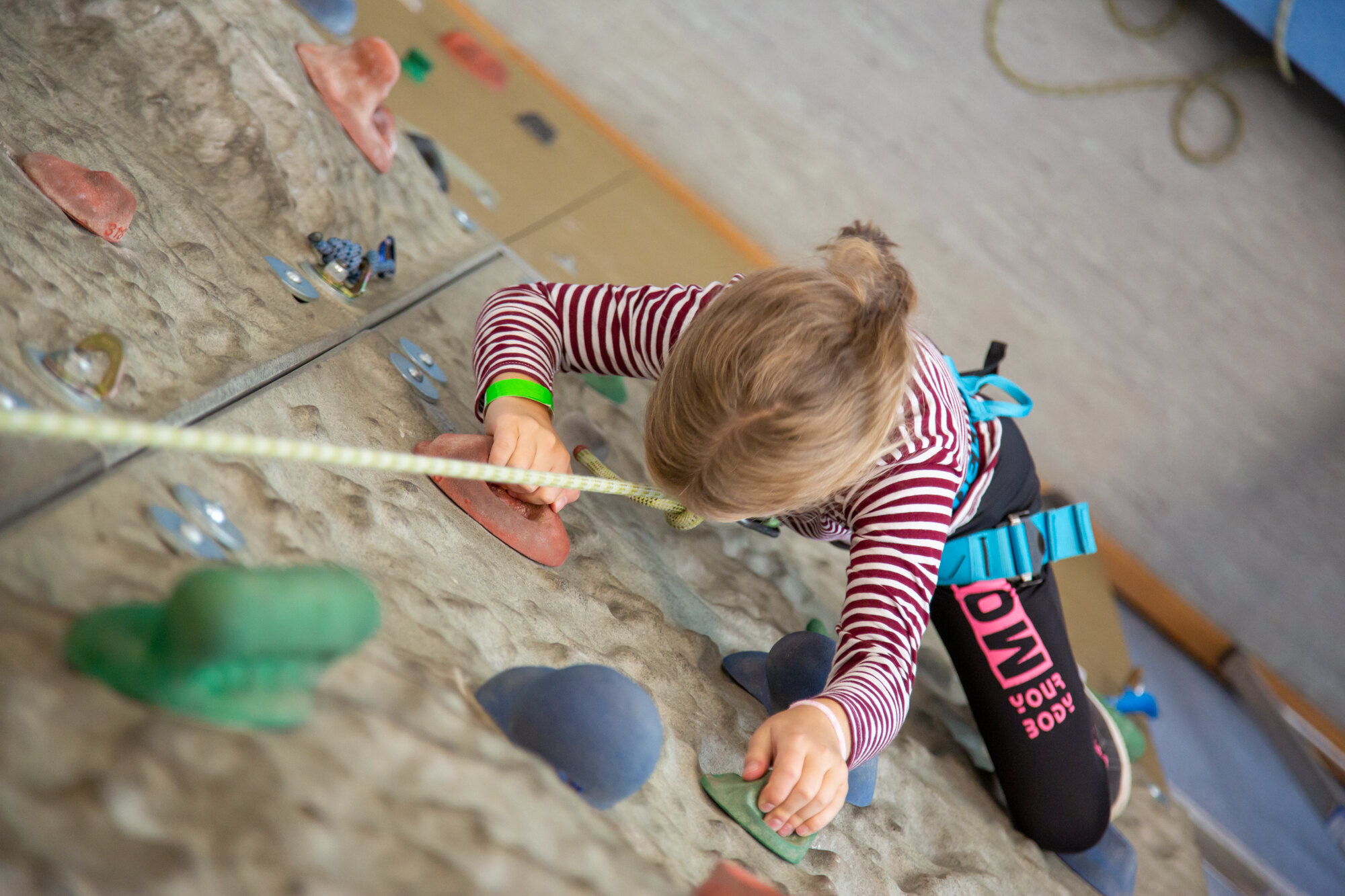 Sportpark Rabenberg: Kletterwand Das Bild zeigt ein Kind beim Klettern an einer Kletterwand. Das Kind ist von oben fotografiert, so dass man es von Kopf bis Fuß sehen kann, während es die Wand erklimmt. Es ist ein junges Mädchen, dessen blondes Haar zu einem kleinen Pferdeschwanz zusammengebunden ist. Sie trägt ein langärmliges rot-weiß gestreiftes Oberteil, schwarze Leggings und einen blauen Klettergurt. Ein Sicherungsseil ist an dem Klettergurt des Kindes befestigt und verläuft nach oben und aus dem Bild heraus.