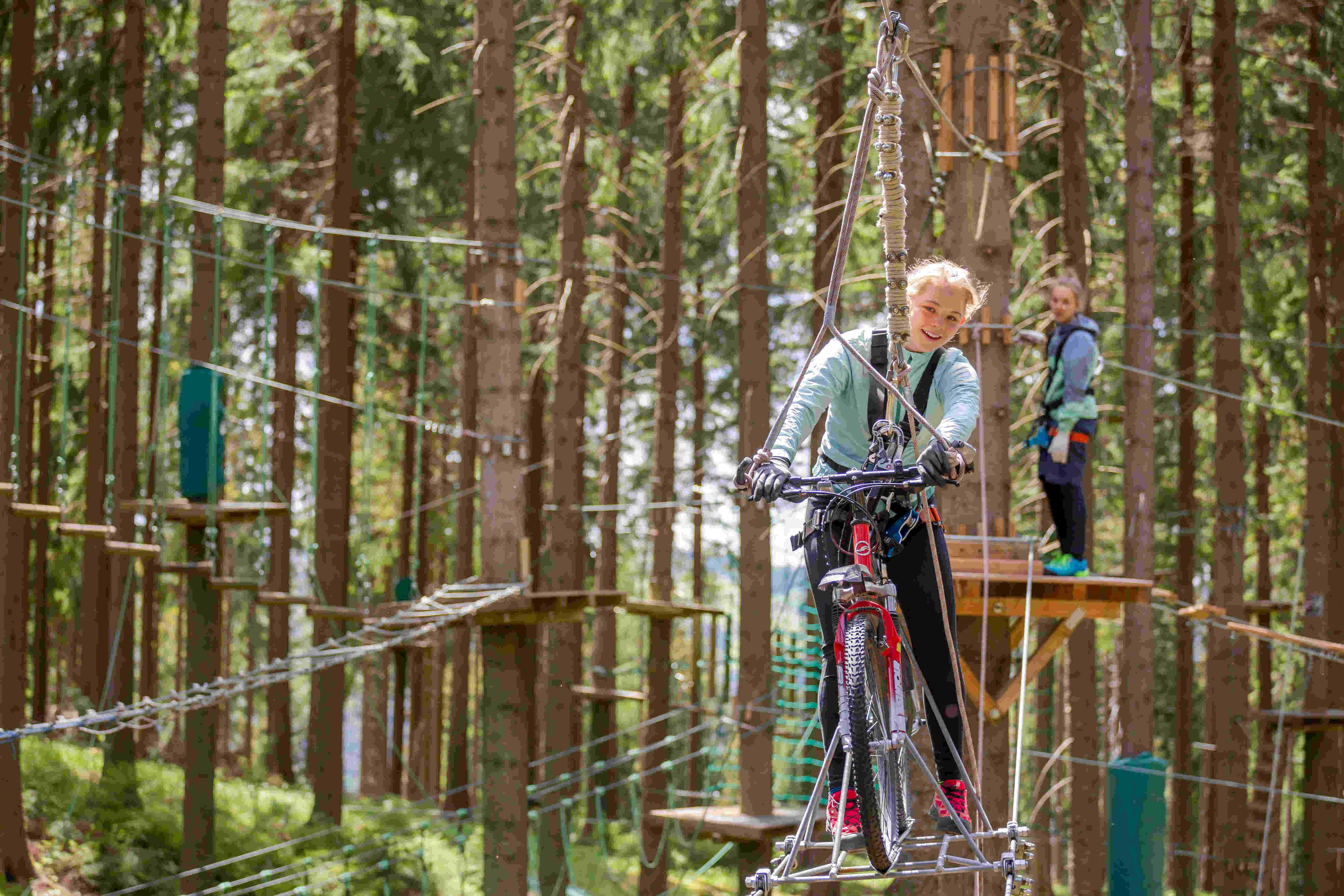 Sportpark Rabenberg: Abenteuerpark Fahrrad Das Bild zeigt eine junge Frau, die mit einem Fahrrad über einen Hochseilgarten fährt. Sie ist in einer grünen Jacke und schwarzen Hosen gekleidet und trägt Handschuhe. Sie hat ein breites Lächeln im Gesicht und schaut in die Kamera. Das Fahrrad ist rot und hat dicke Reifen. Im Hintergrund ist ein anderer junger Mensch zu sehen, der auf einer Plattform steht und ebenfalls einen Gurt trägt. Beide sind von Bäumen umgeben, und es gibt eine Reihe von Hindernissen, die durch den Hochseilgarten führen.