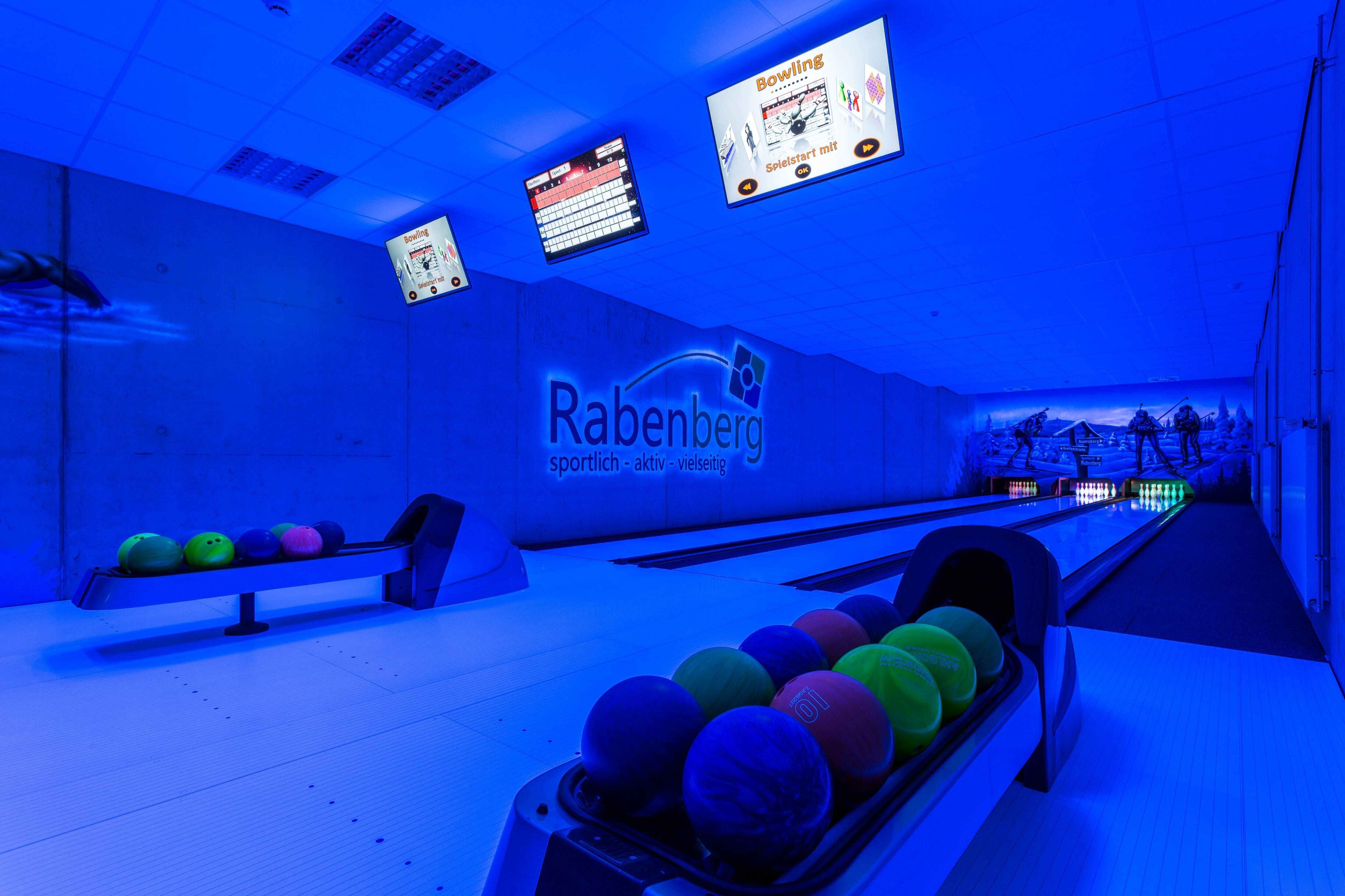 Sportpark Rabenberg: Schwarzlicht Bowlingbahn Das Bild zeigt eine Bowlingbahn in bläulichem Licht. Im Vordergrund befinden sich zwei Kugellager mit farbigen Bowlingkugeln. Am Ende der Bahnen stehen die Kegel, dahinter ist eine Wandmalerei mit Berglandschaft und Skifahrern zu erkennen. Über den Bahnen sind mehrere Monitore an der Decke angebracht, die Informationen über das Bowling anzeigen. Neben der linken Bowlingbahn ist das Logo „Rabenberg“ an der Wand angebracht.