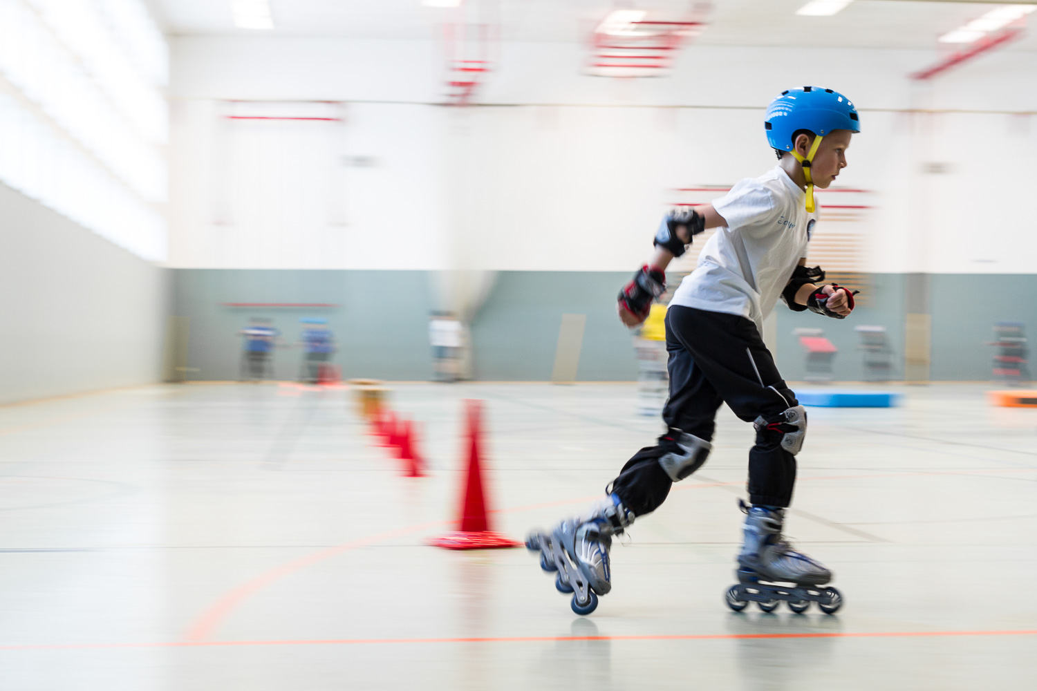 Sportpark Rabenberg: Inlineskating Das Bild zeigt einen Jungen, der auf Inlineskates in einer Halle fährt. Er ist mit einem blauen Helm, Knieschützern, Ellbogenschützern und Handschuhen geschützt. Er trägt ein weißes T-Shirt und eine schwarze Hose. Er fährt mit hoher Geschwindigkeit, was durch die Bewegungsunschärfe im Hintergrund angezeigt wird. Auf dem Boden sind rote Kegel aufgereiht, die wahrscheinlich einen Parcours bilden.