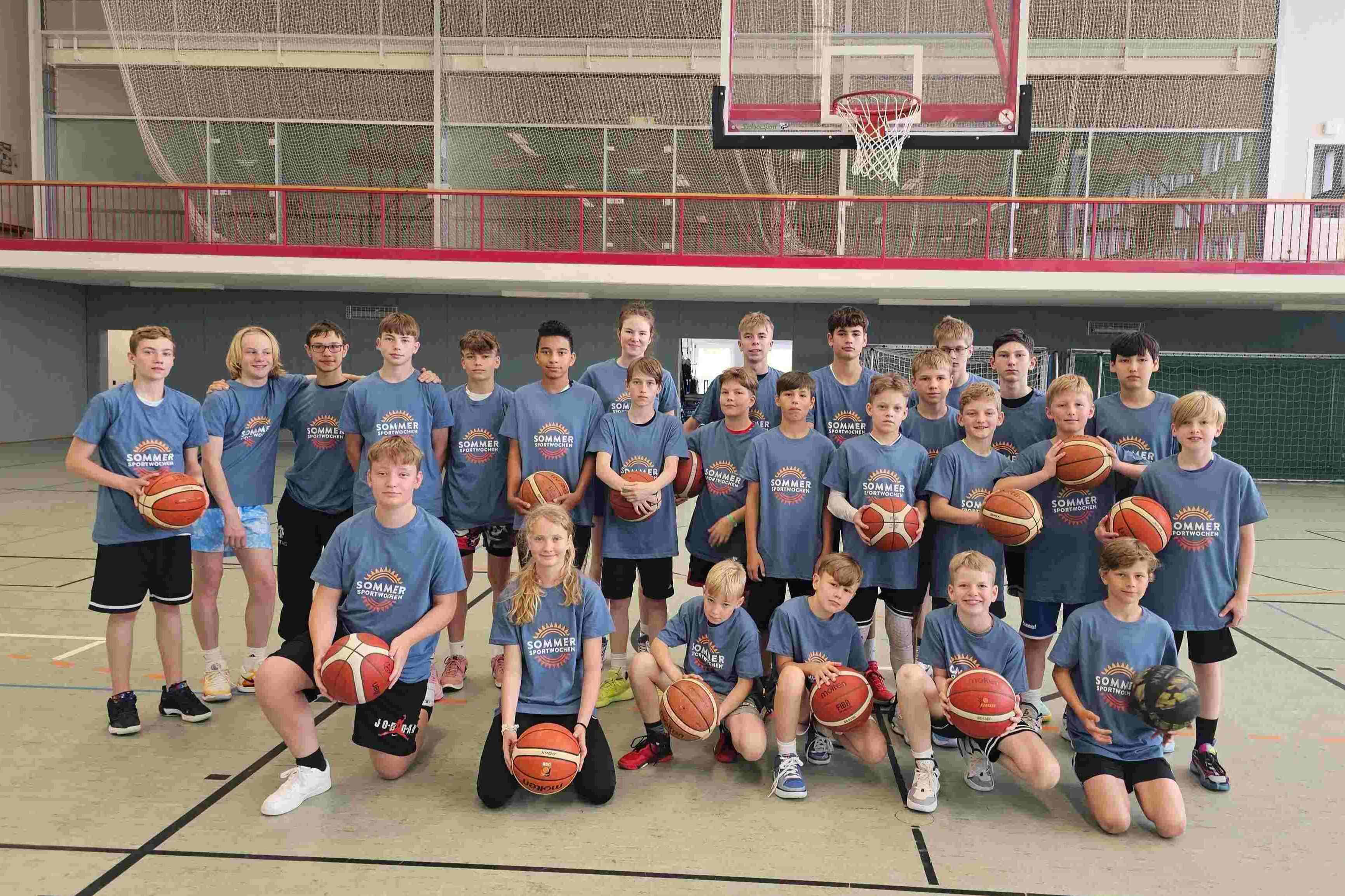 Sportpark Rabenberg: Sommersportwochen Gruppenbild Das Bild zeigt eine Gruppe von Kindern und Jugendlichen, die in einer Sporthalle posieren. Alle tragen blaue T-Shirts mit dem Aufdruck "SOMMERSPORTWOCHEN". Die meisten halten einen Basketball in den Händen. Im Hintergrund ist ein Basketballkorb zu sehen, der an der Wand befestigt ist. Die Gruppe steht in mehreren Reihe, wobei einige Personen knien oder sich in der Hocke befinden. Die Stimmung wirkt locker und fröhlich. Es scheint sich um eine Sportveranstaltung oder ein Trainingscamp zu handeln.