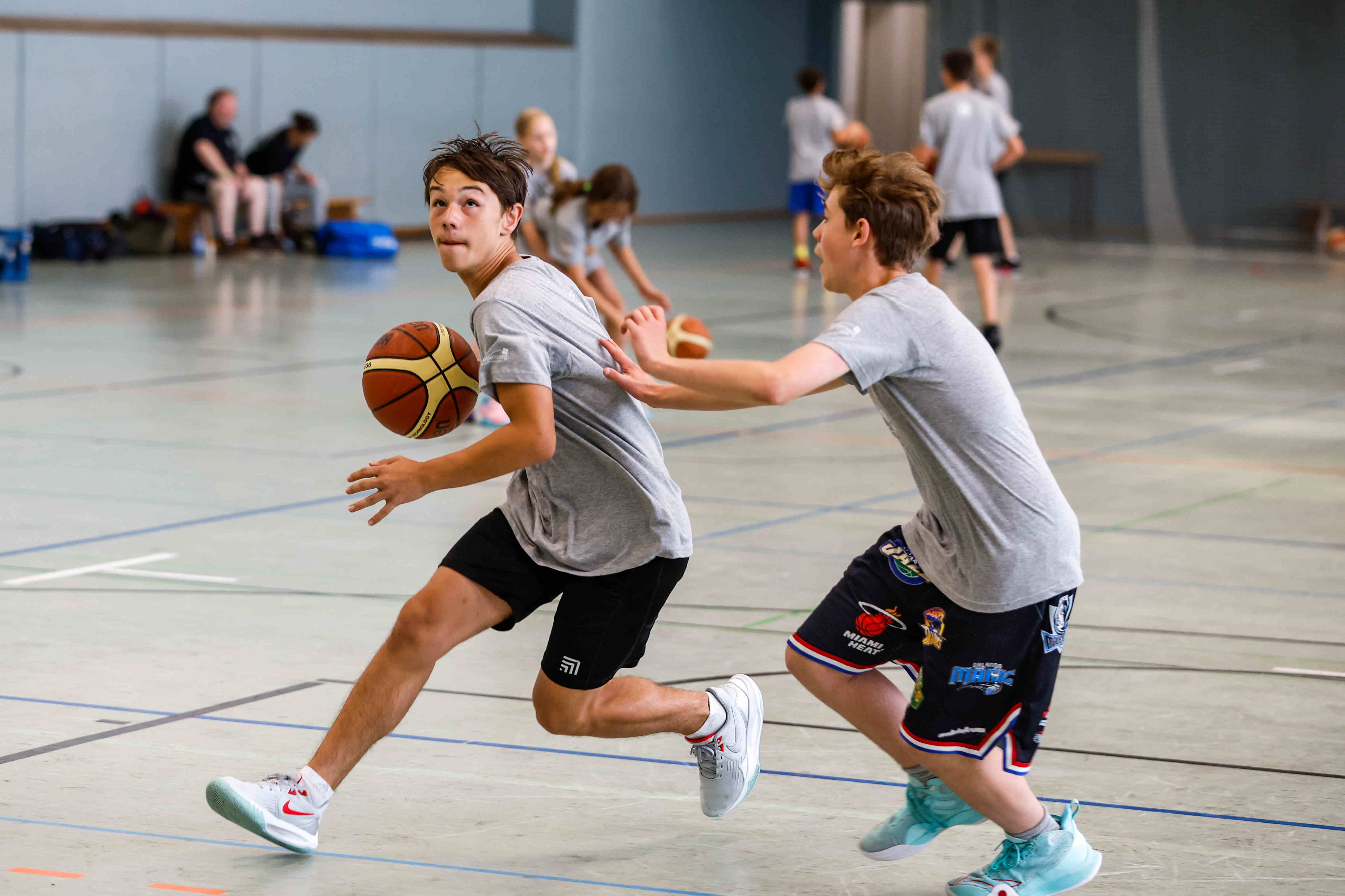 Sportpark Rabenberg: Sommersportwochen Basketball Das Bild zeigt zwei Jungen, die in einer Turnhalle Basketball spielen. Der Junge im Vordergrund dribbelt den Ball mit der rechten Hand und blickt nach links. Er trägt ein graues T-Shirt, schwarze Shorts und weiße Turnschuhe mit roten Akzenten. Der Ball ist orange-braun mit schwarzen Rillen. Der andere Junge versucht, ihm den Ball abzunehmen und streckt seine Hand nach dem ersten Jungen aus. Er trägt ein graues T-Shirt, Basketballshorts mit Teamlogos und hellblaue Turnschuhe. Im Hintergrund sind weitere Personen zu sehen, einige davon scheinen ebenfalls Basketball zu spielen, während andere zuschauen. Sie tragen ähnliche Outfits wie die beiden Jungen im Vordergrund. Auf dem Boden sind Linien für das Spielfeld aufgezeichnet.
