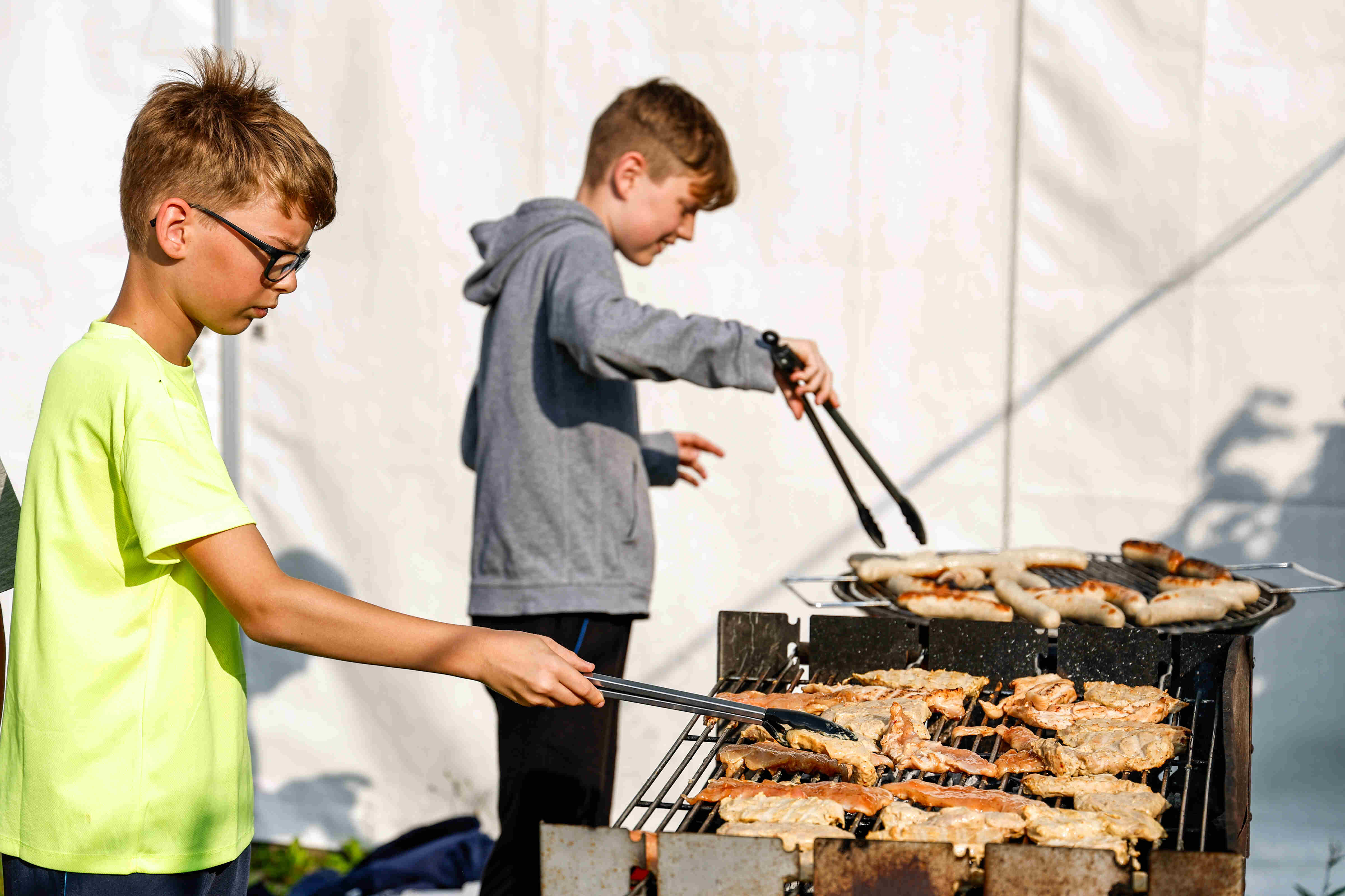 Sportpark Rabenberg: Sommersportwochen Grillabend Das Bild zeigt zwei Jungen, die im Freien grillen. Der Junge auf der linken Seite trägt ein neongelbes T-Shirt und eine Brille. Er hält eine Zange und scheint Fleisch auf dem Grill zu wenden. Der andere Junge, der sich im Hintergrund befindet, trägt einen grauen Kapuzenpullover und scheint ebenfalls mit der Grillzange zu hantieren. Auf dem Grill liegen Würste und Fleischstücke. Der Hintergrund ist ein weißes Zelt. Die Szene wirkt sommerlich und entspannt.