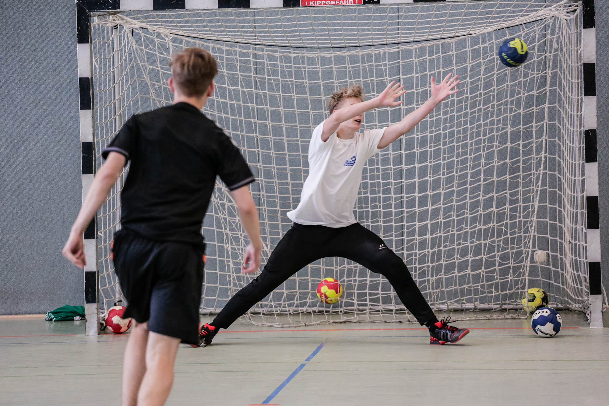 Sportpark Rabenberg: Sommersportwochen Handball Das Bild zeigt eine Szene aus einem Handballspiel. Im Vordergrund sind zwei Jungen zu sehen. Einer der Jungen steht mit dem Rücken zur Kamera und trägt ein schwarzes T-Shirt und eine schwarze Hose. Er scheint gerade einen Wurf auszuführen. Der andere Junge steht im Tor und versucht, den Ball zu fangen. Er trägt ein weißes T-Shirt und eine schwarze Hose. Er hat die Arme weit geöffnet und versucht, den Ball abzuwehren. Der Ball ist gerade im Flug und nähert sich dem Tor. Im Hintergrund ist ein Handballtor zu sehen, das aus einem Netz und einem Rahmen besteht. Auf dem Boden liegen weitere Bälle. Die Halle hat graue Wände.