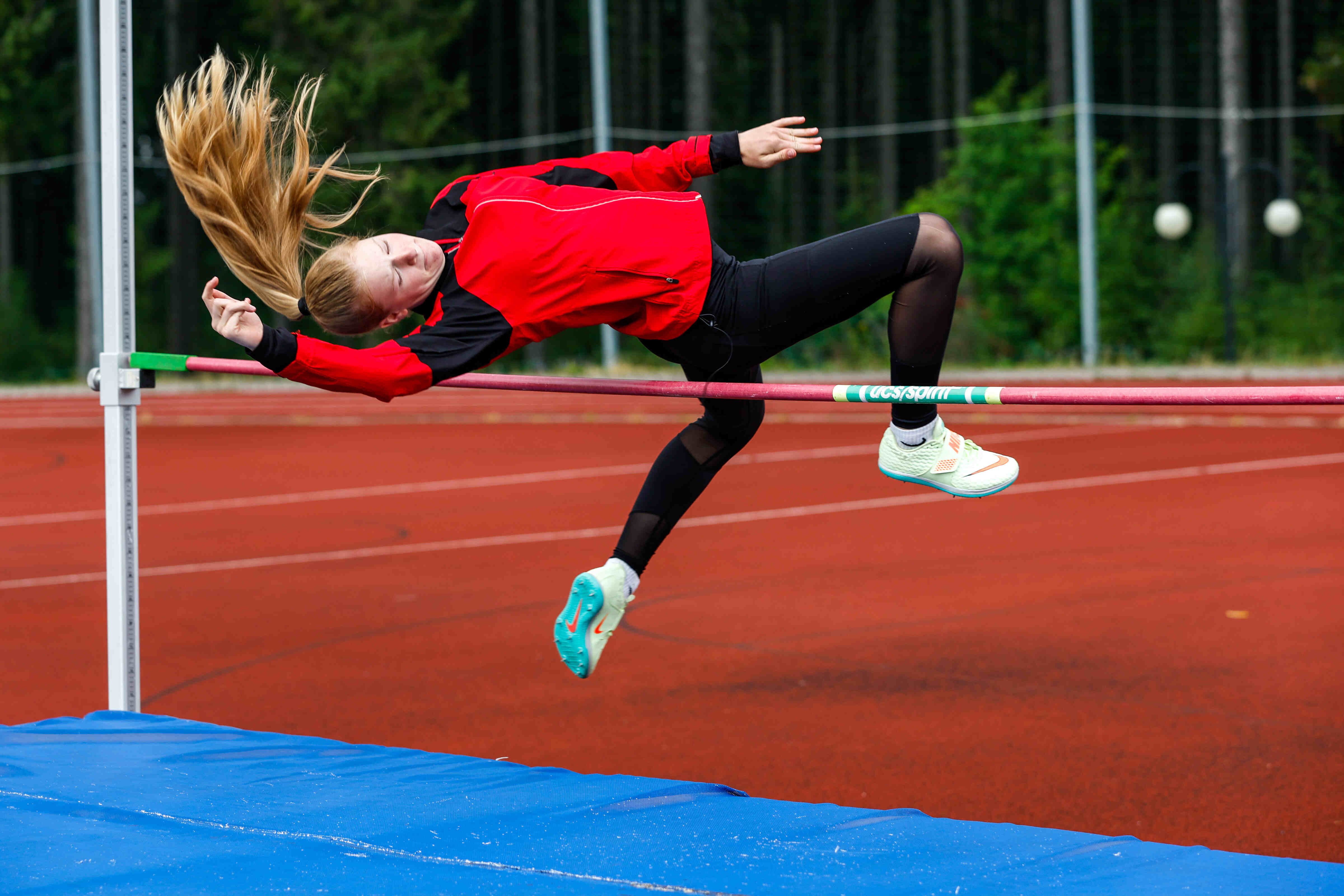 Sportpark Rabenberg: Sommersportwochen Hochsprung Das Bild zeigt ein Mädchen, das Hochsprung macht. Es ist in der Luft und versucht, über die Latte zu springen. Sie trägt eine rote Jacke, schwarze Leggings und Laufschuhe. Ihre Haare sind blond und fliegen in der Luft. Die Latte ist rot und grün. Das Bild wird auf einer roten Sportbahn vor einem grünen Hintergrund aufgenommen. Unter der Latte befindet sich eine blaue Matte.