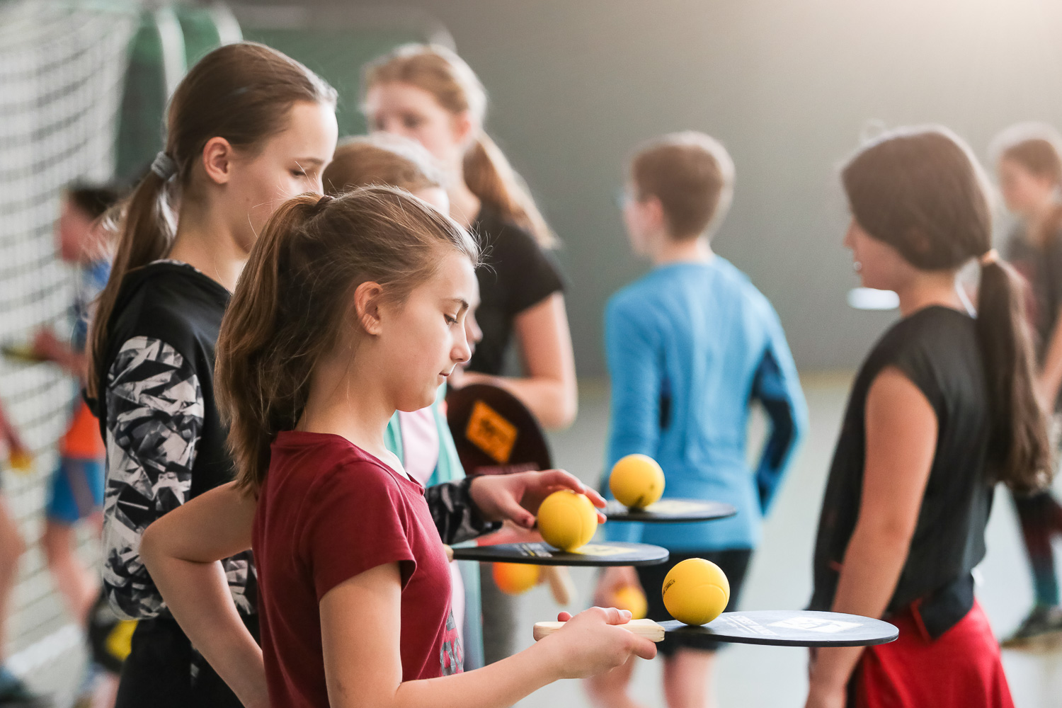 Sportpark Rabenberg: Sommersportwochen Sportspiele Das Bild zeigt eine Gruppe von Kindern in einer Sporthalle. Sie scheinen gerade eine Sportart zu spielen, bei der sie kleine gelbe Bälle mit Schlägern schlagen. Im Vordergrund stehen mehrere Mädchen die jeweils einen Ball auf dem Schläger balancieren. Eines der Mädchen in einem roten T-Shirt hält einen Schläger in der Hand und legt gerade einen gelben Ball auf den Schläger. Andere Mädchen halten ebenfalls Schläger und Bälle. Im Hintergrund sind weitere Kinder zu sehen, die in verschiedene Richtungen schauen oder sich bewegen.