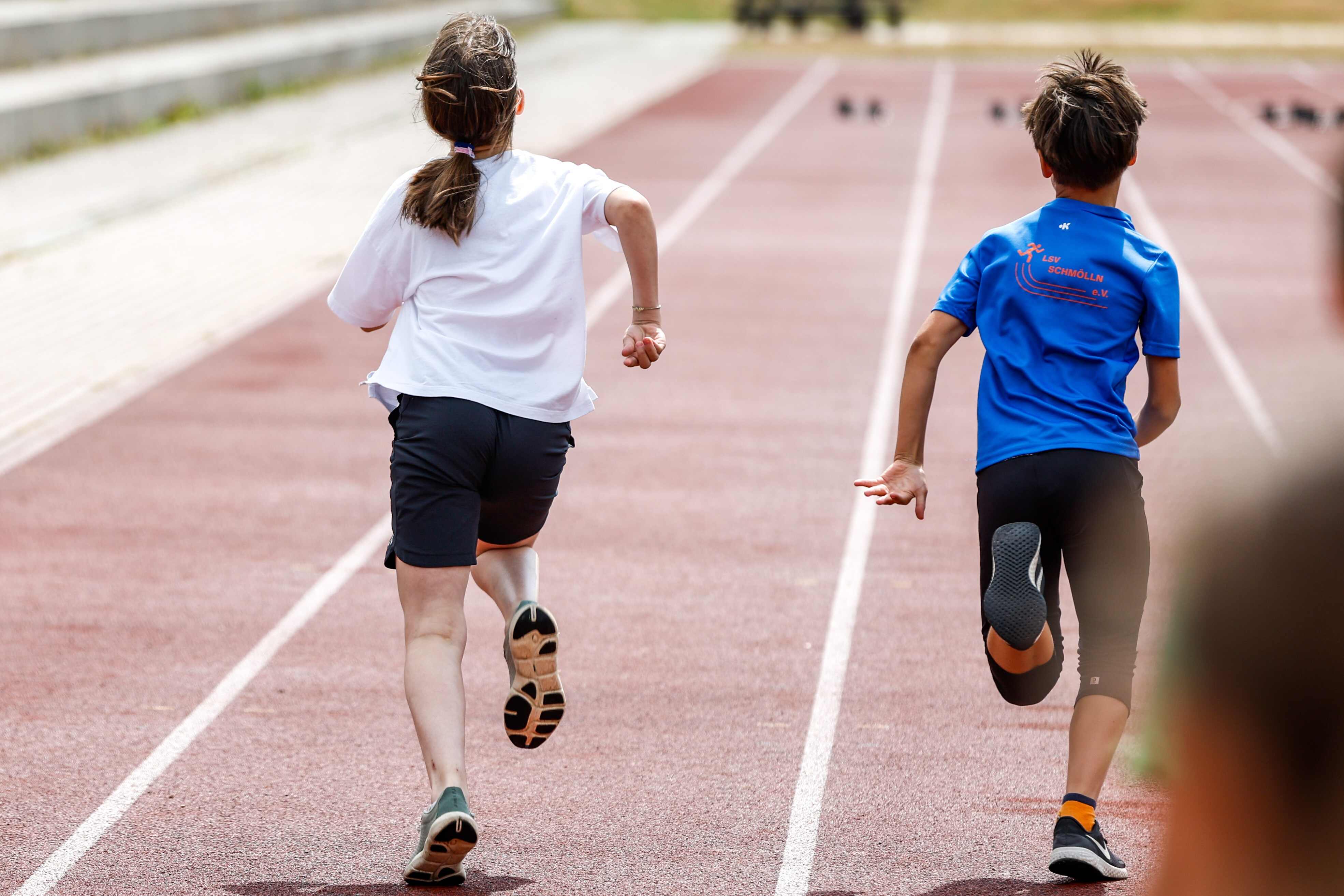 Sportpark Rabenberg: Sommersportwochen Sprint Das Bild zeigt zwei Kinder, die auf einer roten Laufbahn rennen. Beide sind von hinten zu sehen. Das Mädchen, das links auf dem Bild rennt, trägt ein weißes T-Shirt und schwarze Shorts. Sie hat ihr Haar zu einem Pferdeschwanz zusammengebunden. Ihre Arme sind angewinkelt. Der Junge, der rechts auf dem Bild rennt, trägt ein blaues T-Shirt mit einem Logo auf der Rückseite. Er trägt schwarze Shorts und schwarze Schuhe mit orangefarbenen Socken. Seine Arme sind angewinkelt. Die Laufbahn hat weiße Markierungen.