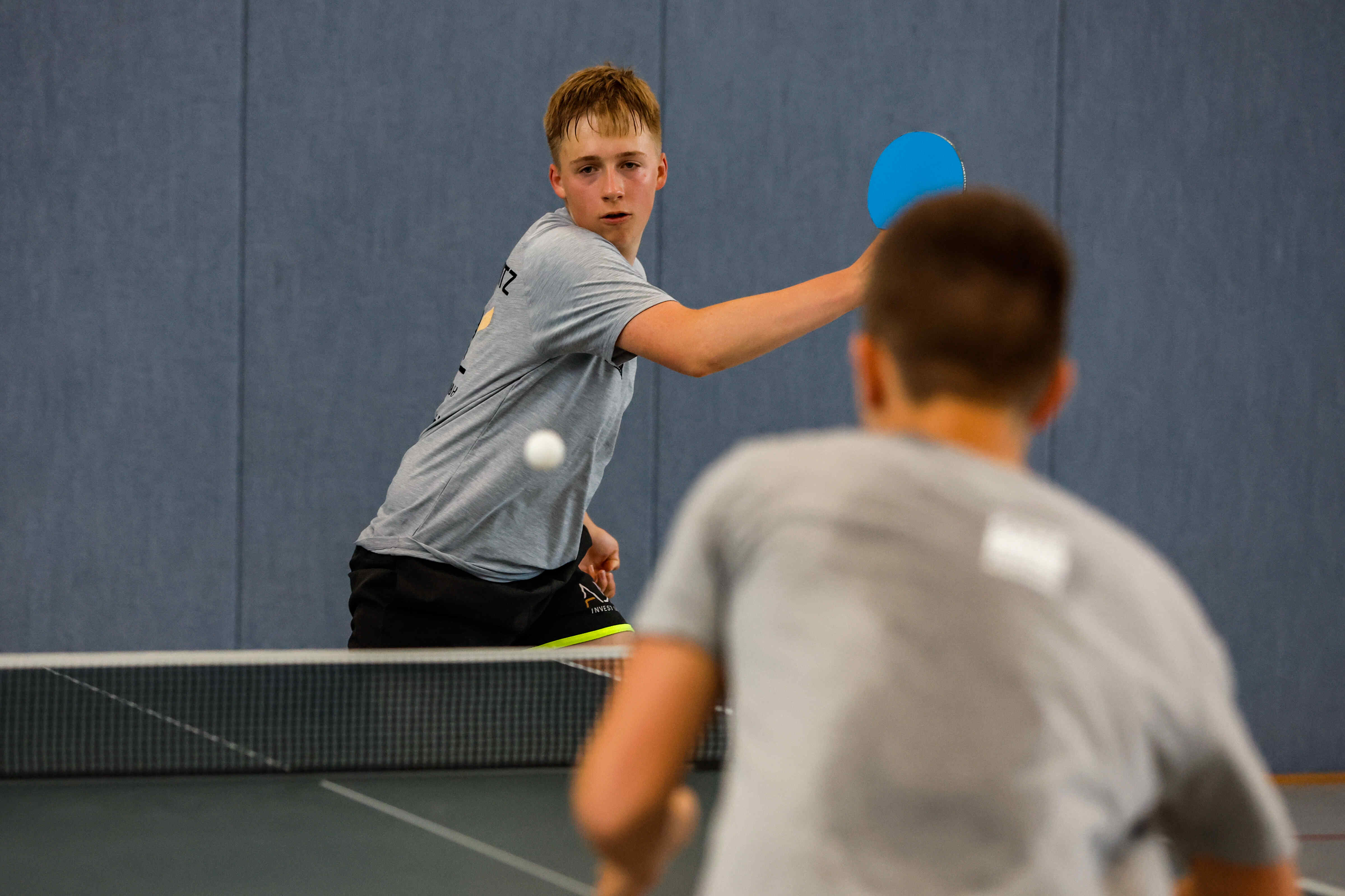 Sportpark Rabenberg: Sommersportwochen Tischtennis Das Foto zeigt zwei Jungen, die Tischtennis spielen. Der Junge im Vordergrund ist unscharf und von hinten zu sehen. Er trägt ein graues T-Shirt. Der andere Junge, der im Fokus steht, ist auch mit einem grauen T-Shirt bekleidet und schaut in Richtung der Kamera. Er hat einen blonden Haarschnitt. Er hält einen blauen Schläger in der Hand und schlägt gerade den Ball. Der Ball ist auch im Fokus und befindet sich in der Mitte des Bildes. Im Hintergrund befindet sich eine blaue Wand und ein Tischtennisnetz.