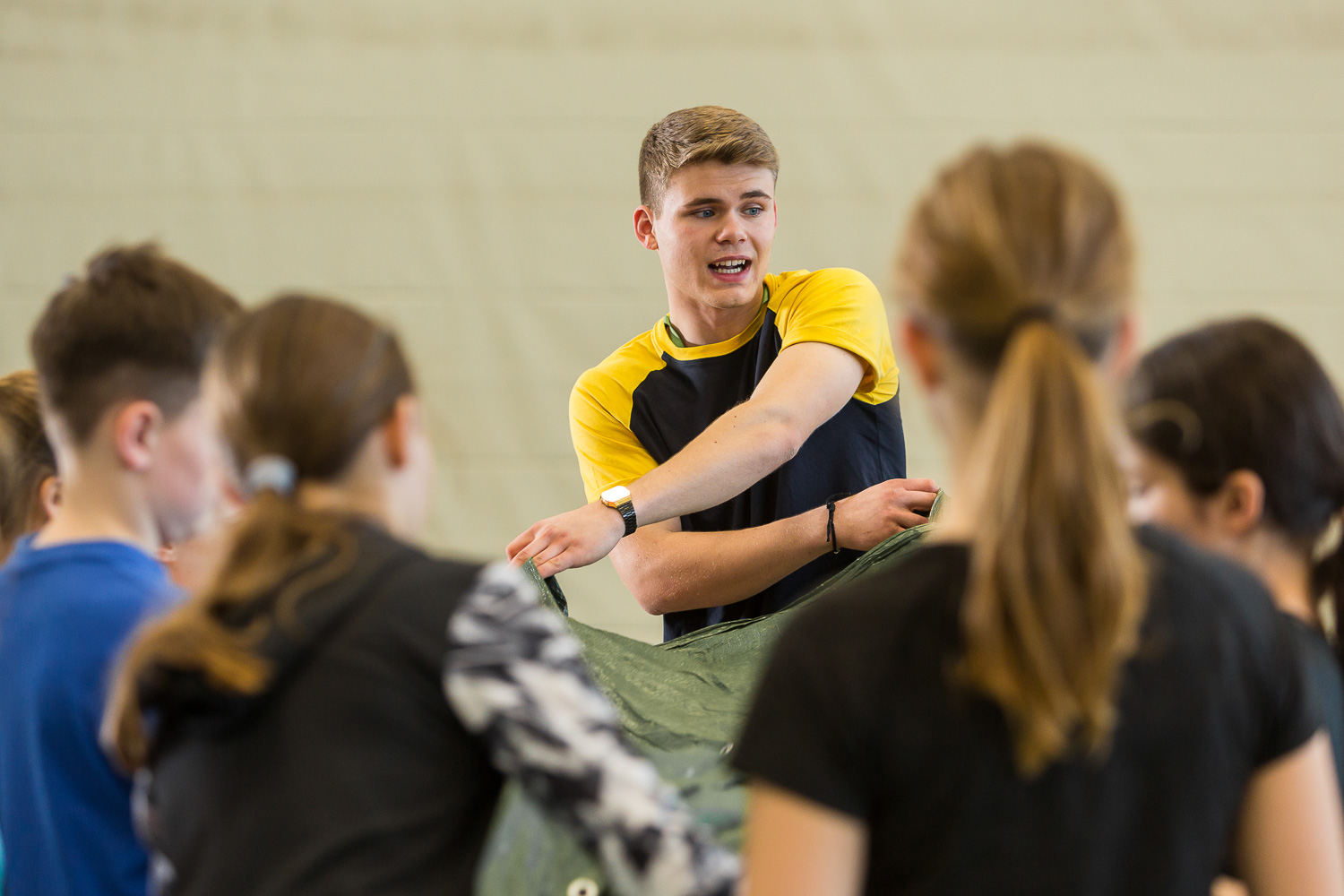 Sportpark Rabenberg: Sommersportwochen Trainer mit Kindergruppe Das Bild zeigt eine Gruppe von Menschen, die in einem hellen Raum zusammenkommen. Im Mittelpunkt steht ein junger Mann, der sich anscheinend an eine Gruppe von Kindern wendet, die ihm gegenüberstehen. Er trägt ein gelb-schwarzes T-Shirt und eine Armbanduhr. Er hält eine grüne Plane in seinen Händen. Die Kinder um ihn herum sind nicht im Detail zu erkennen, aber es scheint, dass sie ihm zuhören.